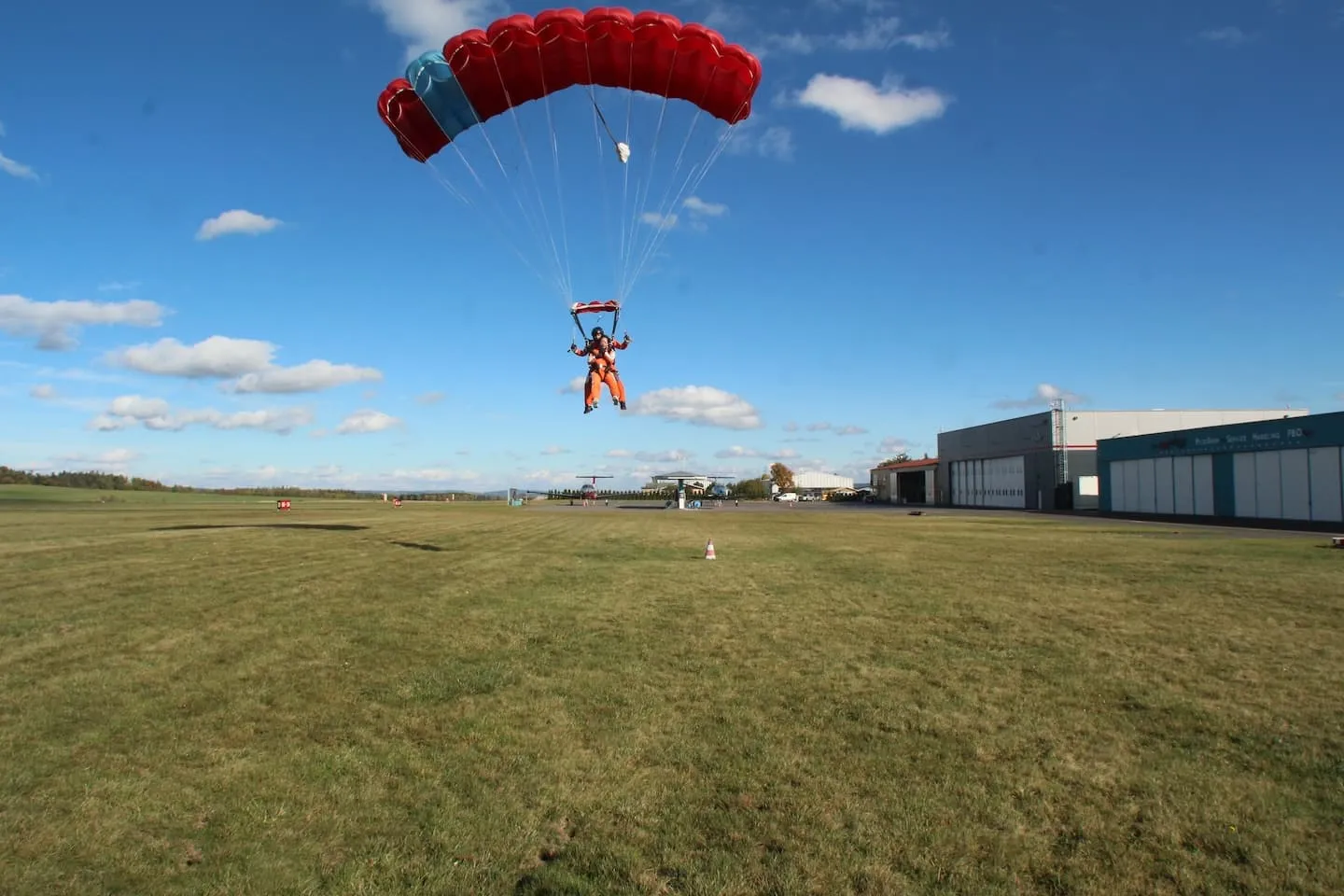 Instructor tándem y pasajero bajo una campana roja, a punto de aterrizar en la hierba del aeródromo, con hangares a la derecha