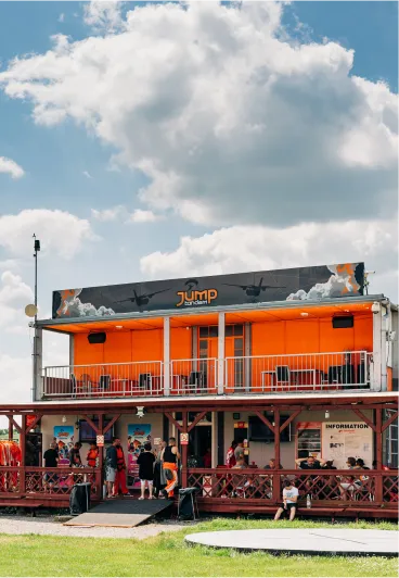 Edificio naranja de JUMP-TANDEM en el aeródromo de Prostějov con terraza en la planta alta y visitantes en el porche de madera