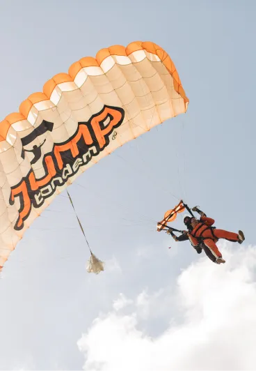 Instructor tándem y pasajero bajo un paracaídas naranja y blanco abierto con el logotipo de JUMP-TANDEM, vistos desde abajo
