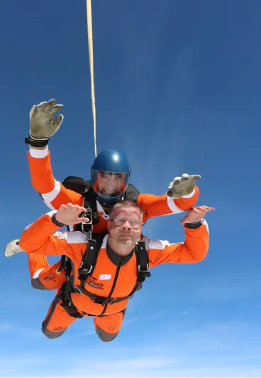 Pasajero con gafas y mono naranja con un instructor tándem con casco azul en caída libre en un cielo azul despejado