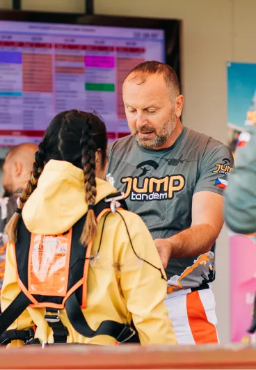 Instructor tándem con camiseta JUMP-TANDEM revisa el arnés de una pasajera con sudadera amarilla y dos trenzas