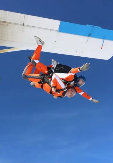 Instructor tándem y pasajero con monos naranjas justo tras la salida bajo el ala blanca y azul del avión