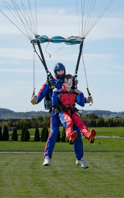 Aterrizaje en tándem: el instructor tándem con mono azul toca la hierba y el pasajero con mono rojo lleva las piernas levantadas