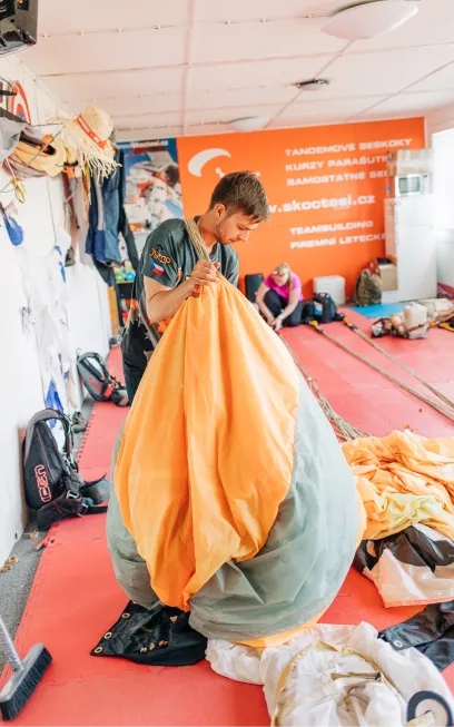 Un plegador dobla un paracaídas naranja y gris en la sala de plegado con moqueta roja