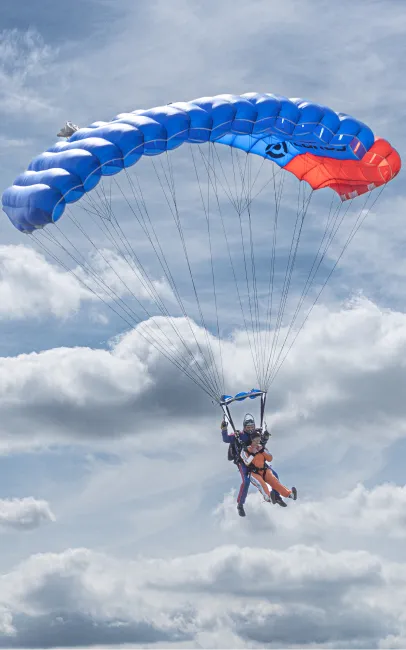 Instructor tándem y pasajero vuelan bajo un paracaídas azul y rojo abierto con nubes de fondo