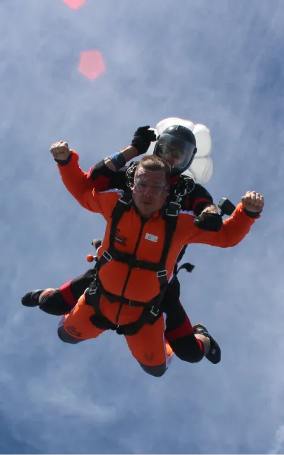 Pasajero con mono naranja y puños cerrados e instructor tándem con casco negro en caída libre, con el paracaídas estabilizador detrás