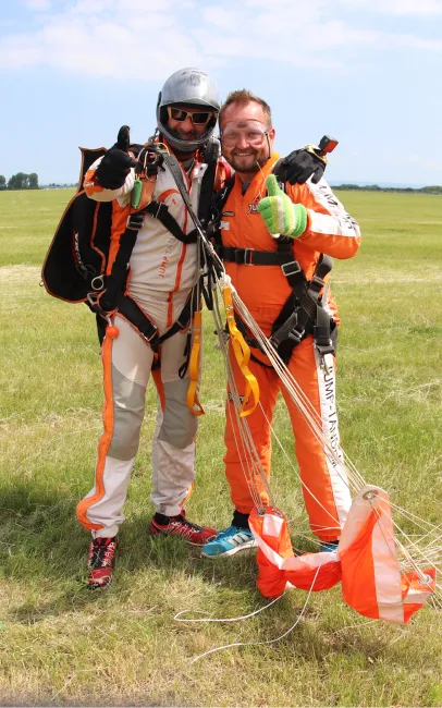 Instructor tándem con casco plateado y pasajero con mono naranja levantan el pulgar tras aterrizar, con las líneas a sus pies