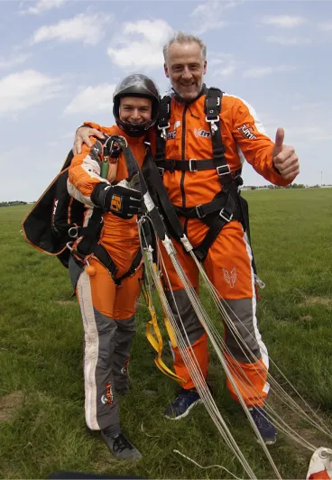Instructor tándem con casco negro y pasajero sonriente con mono naranja abrazados por los hombros tras el aterrizaje