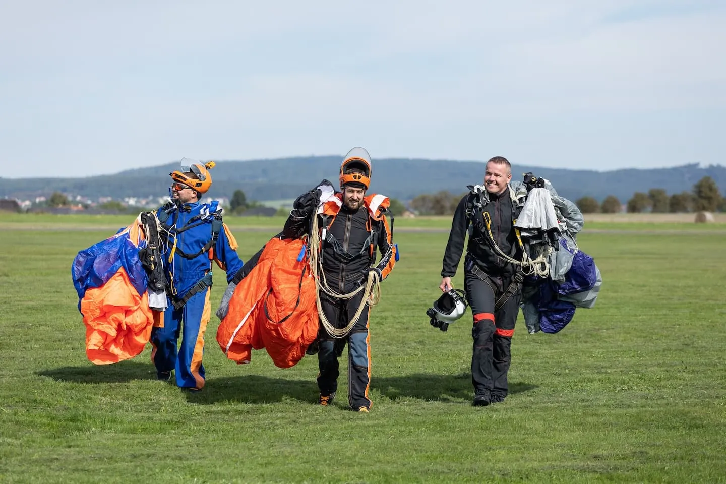 Příbram airport: three skydivers walking across the grass after landing, carrying their bundled parachutes