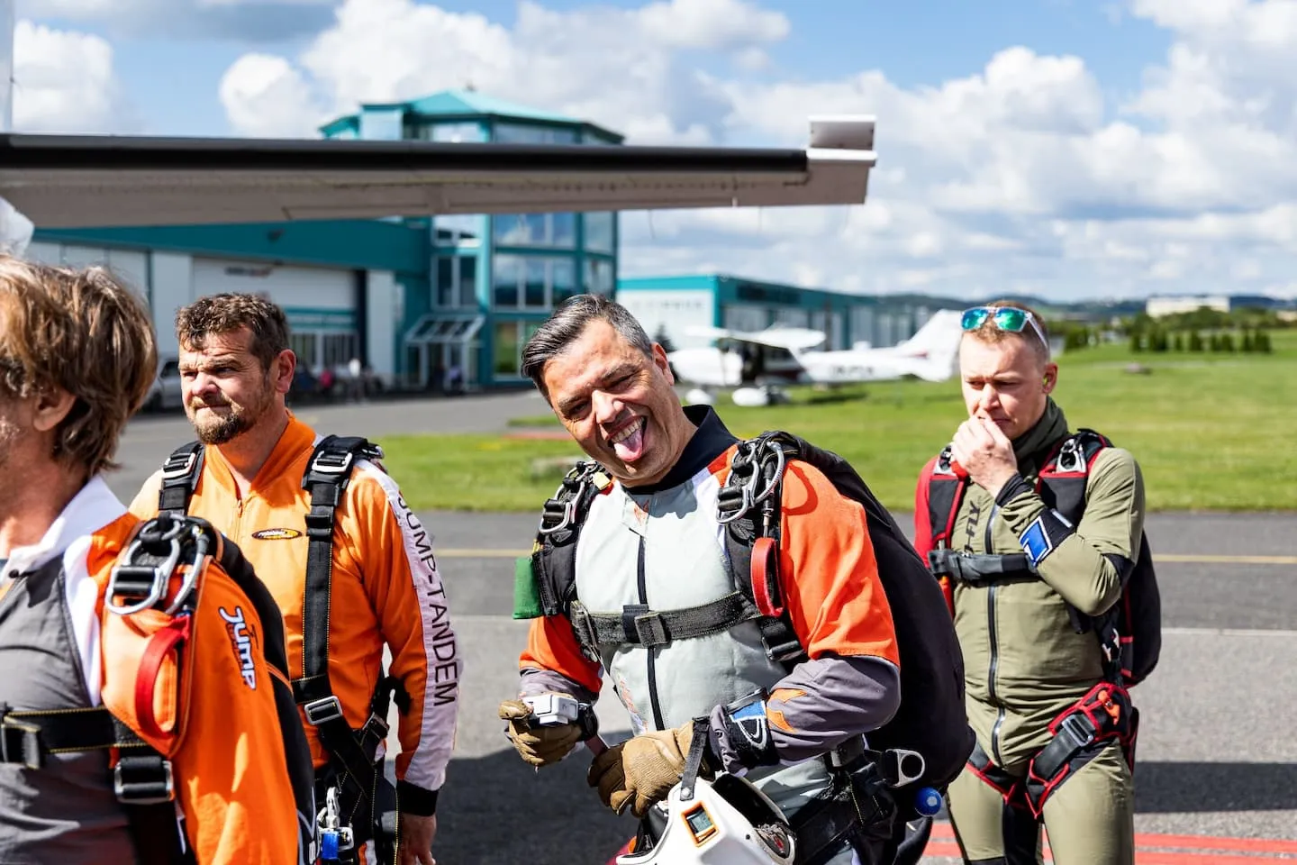 Příbram airport: tandem instructors wearing rigs on the apron, one laughing with his tongue out and holding a camera helmet