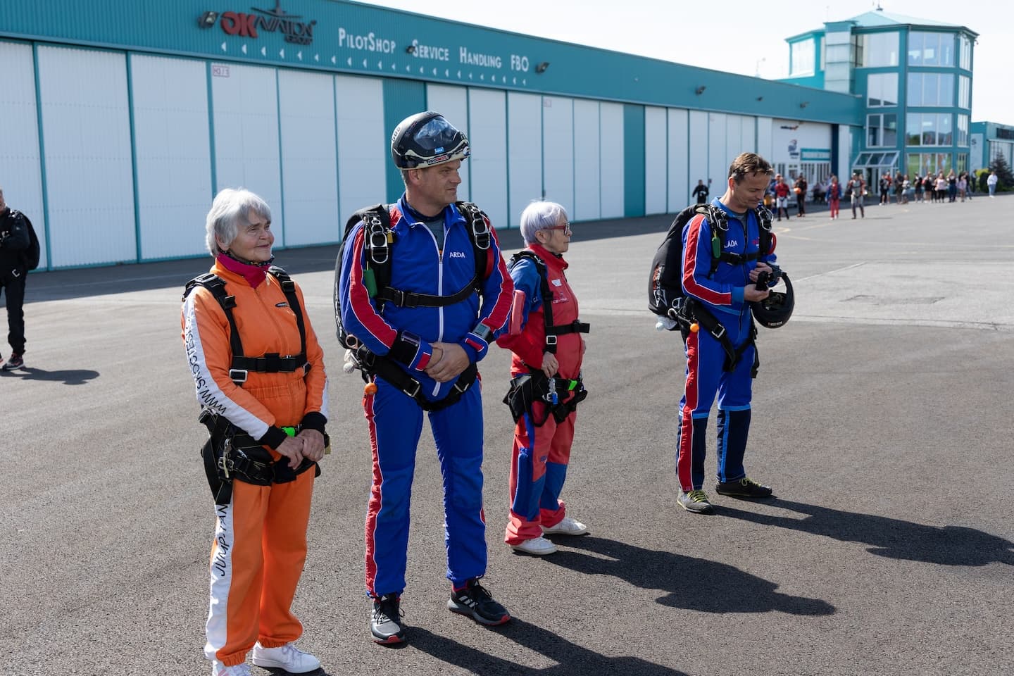 Příbram airport: two passengers in harnesses and two tandem instructors in blue jumpsuits on the apron in front of a hangar