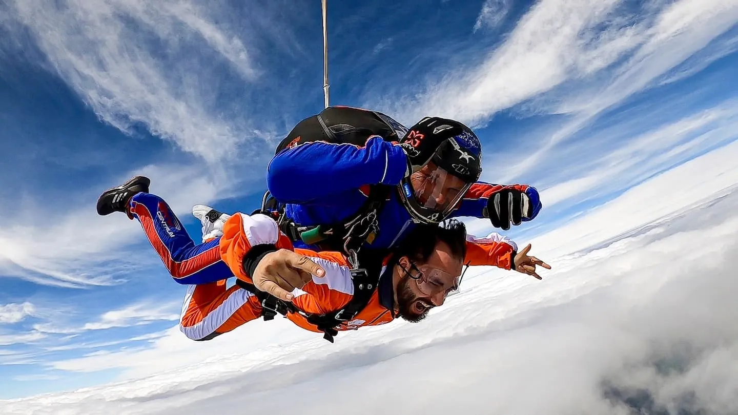 Tandem instructor in a blue jumpsuit and passenger in goggles pointing at the camera, in freefall above the clouds