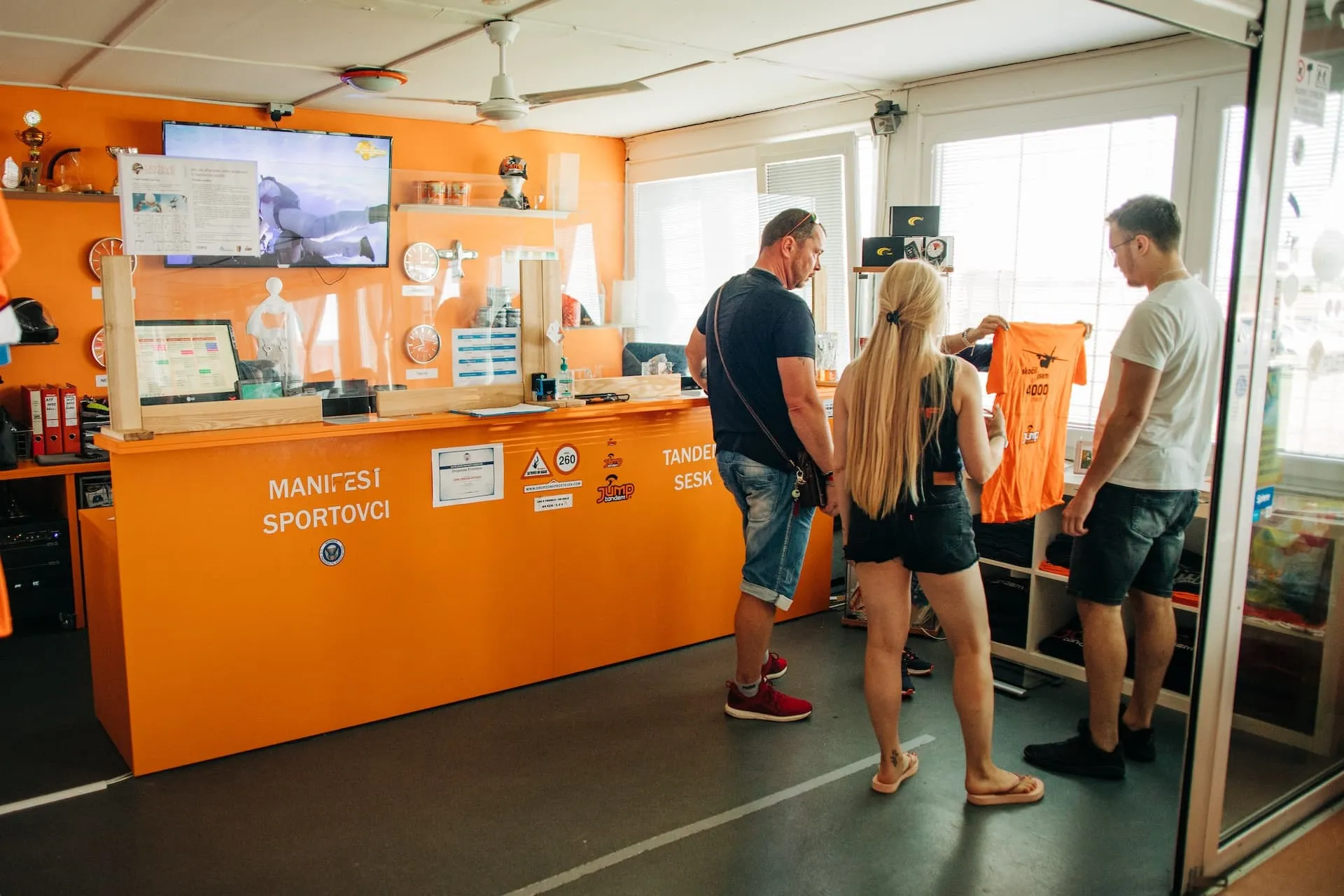 Aeródromo de Prostějov: recepción naranja con el rótulo Manifest sportovci, tres visitantes miran una camiseta naranja