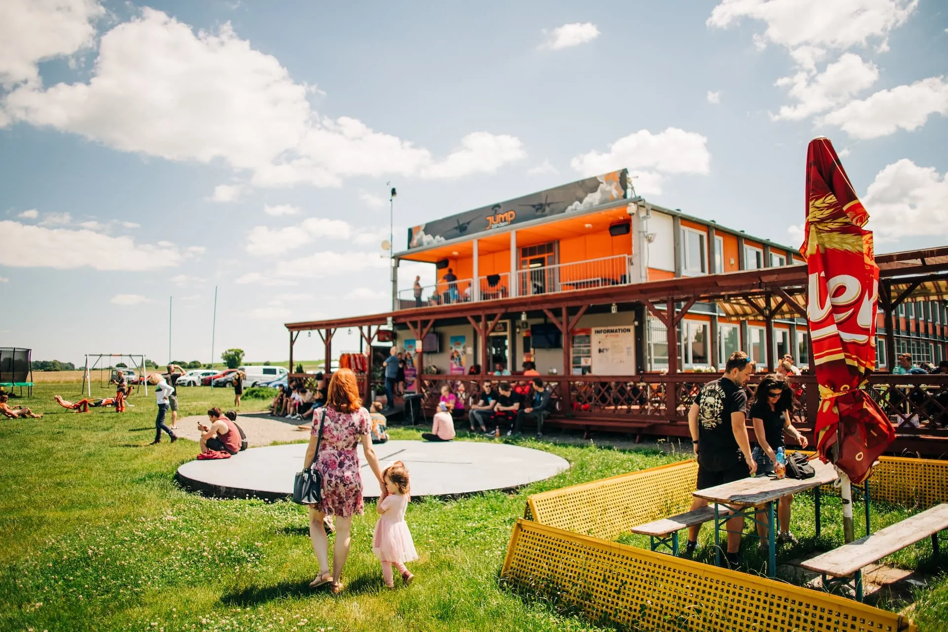 Prostějov airport: a woman and a little girl walking across the grass towards the orange building, visitors sitting around