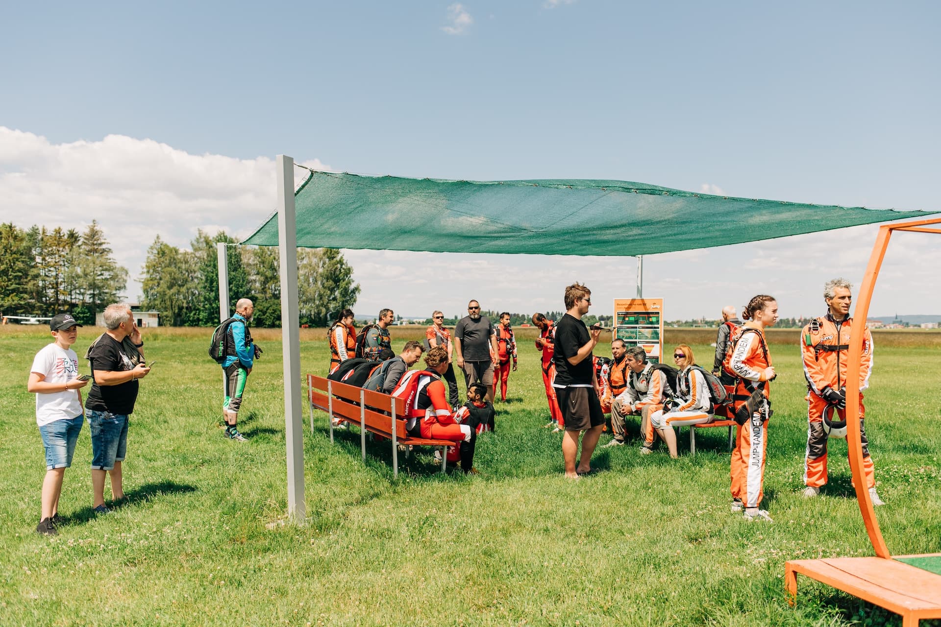 Aeródromo de Prostějov: paracaidistas con monos naranjas y visitantes esperan en bancos bajo un toldo verde sobre la hierba