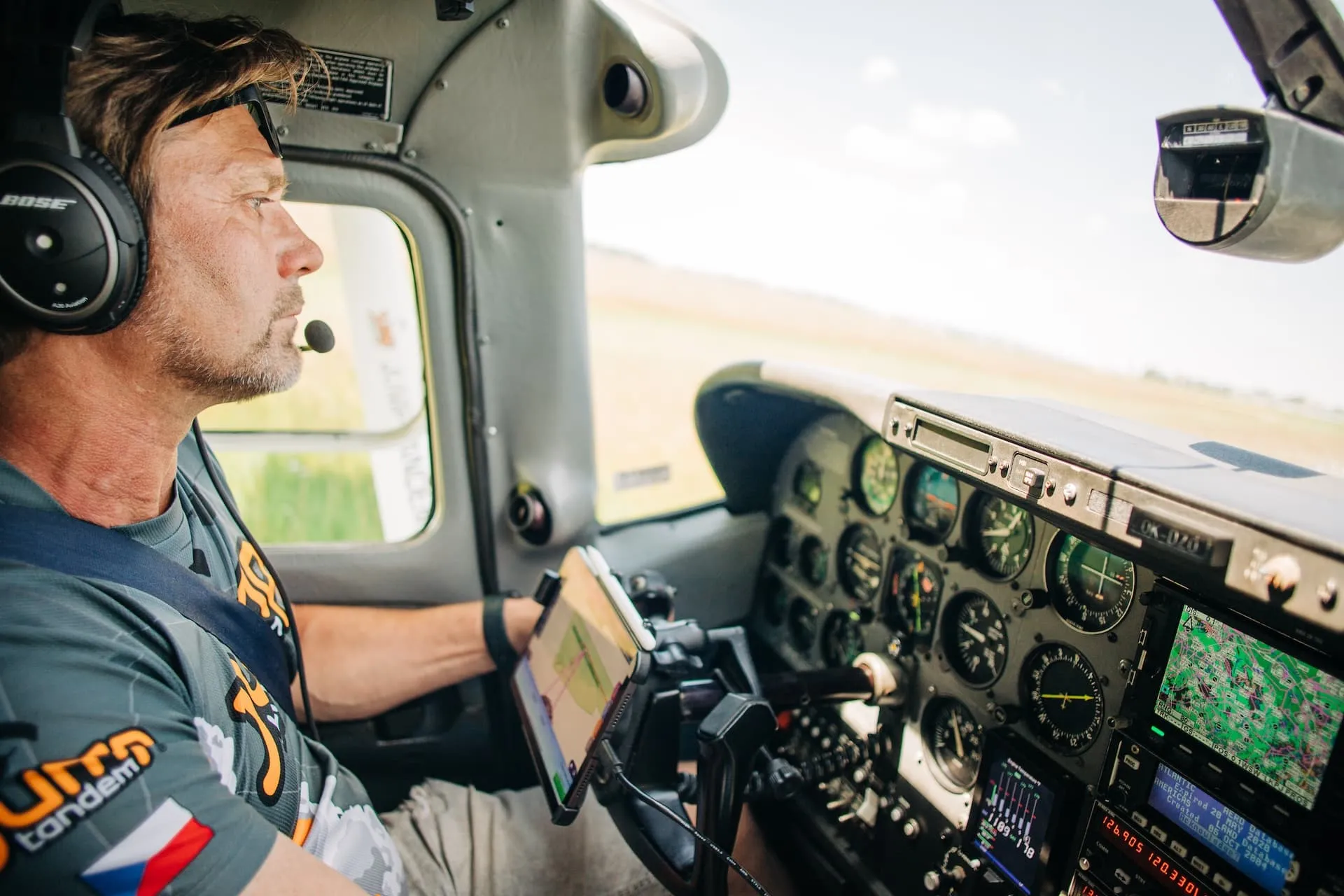 Martin Dlouhý in a JUMP-TANDEM shirt flying a small aircraft, wearing a headset, with a tablet showing a map in front of him