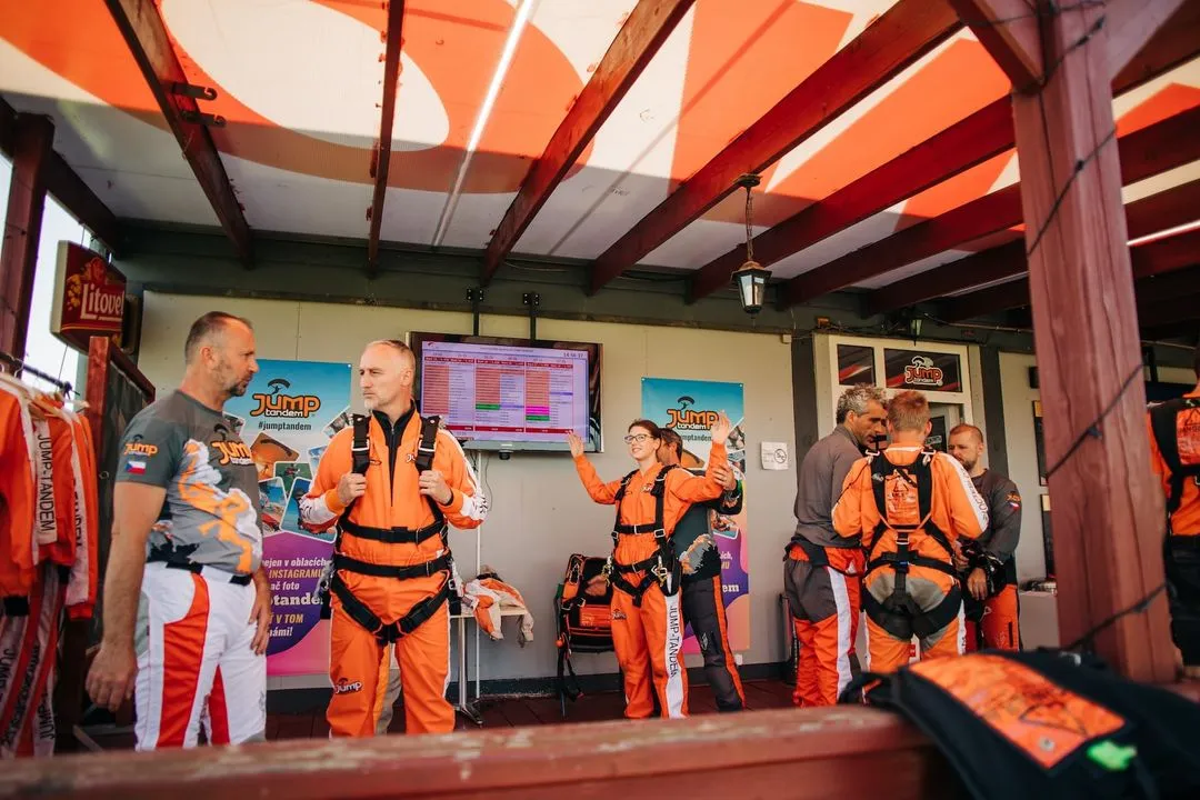Instrucciones antes del salto: pasajeros con monos naranjas y arneses bajo una pérgola, una mujer practica con los brazos en alto