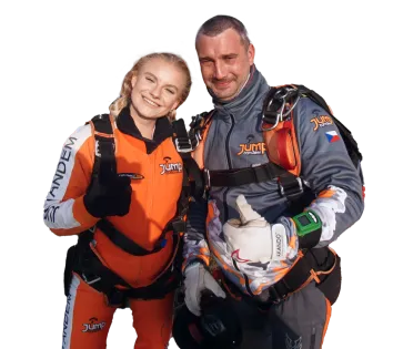 Un instructor tándem con mono gris levanta el pulgar junto a una pasajera sonriente con mono naranja Jump