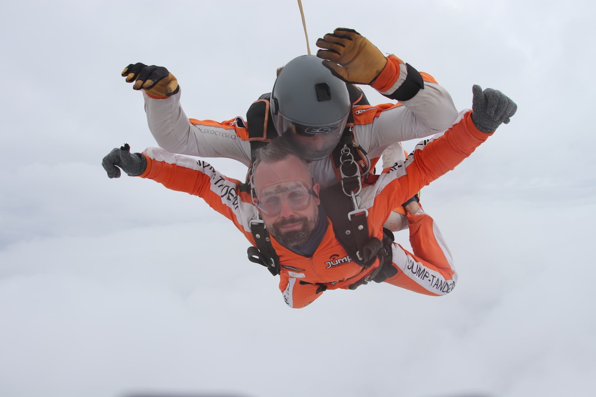 Caída libre sobre un manto de nubes blancas: pasajero con mono naranja y brazos abiertos e instructor tándem con casco gris