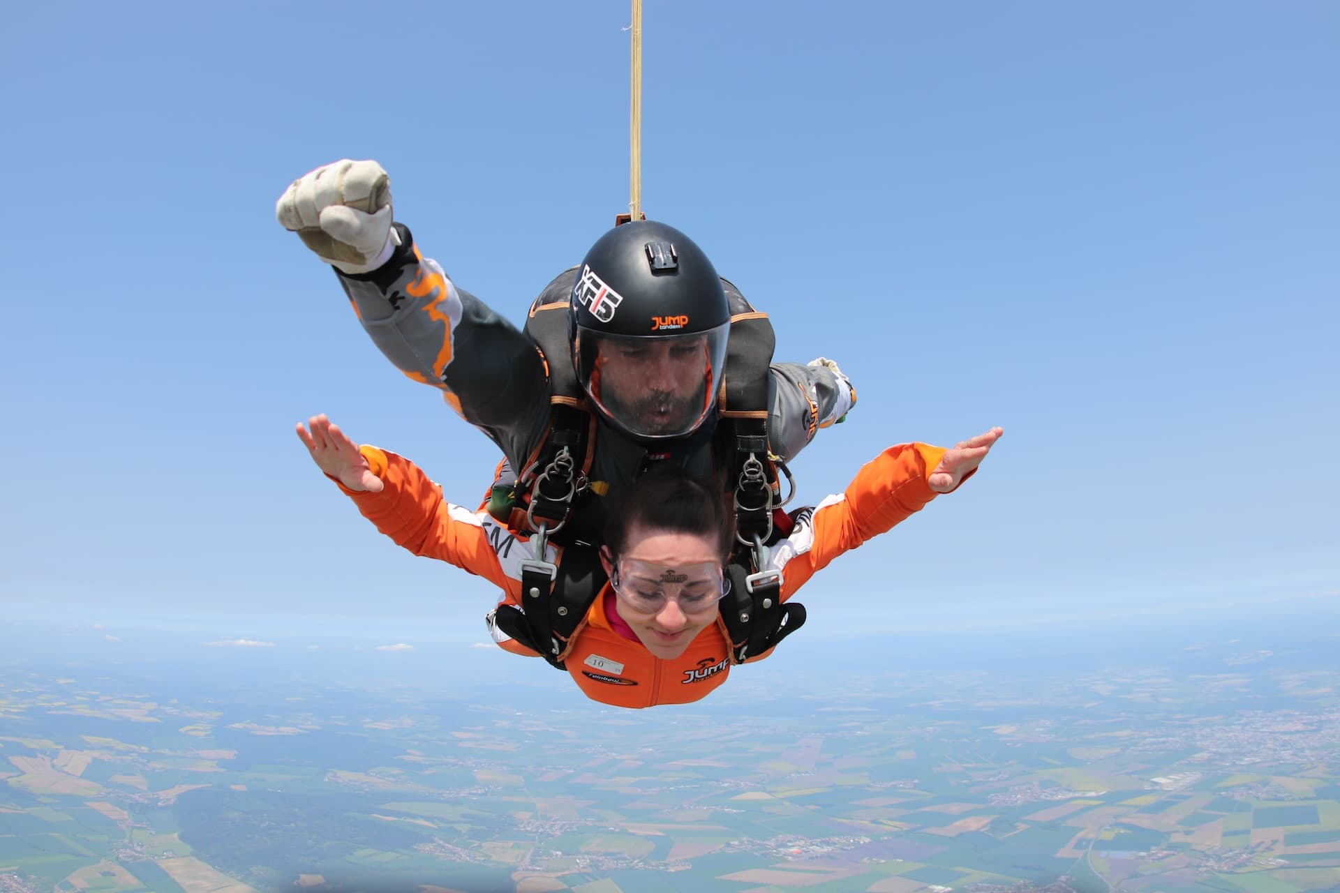 Caída libre: una pasajera con mono naranja y los brazos abiertos, con un instructor tándem de gris y casco negro sobre un paisaje brumoso