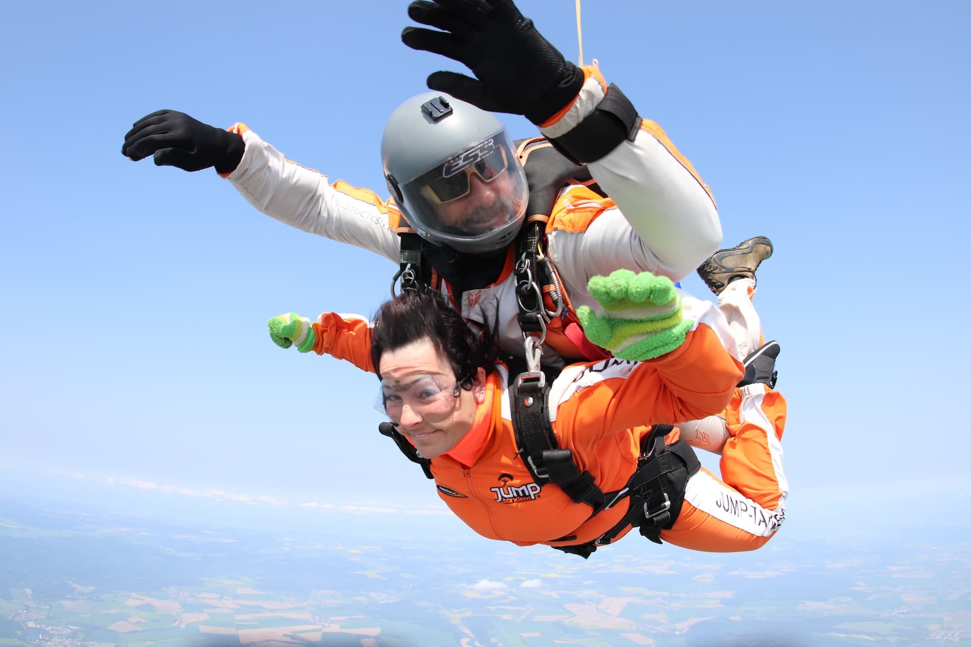 Caída libre: pasajera sonriente con mono naranja y guantes verdes; el instructor tándem con casco gris saluda sobre su cabeza