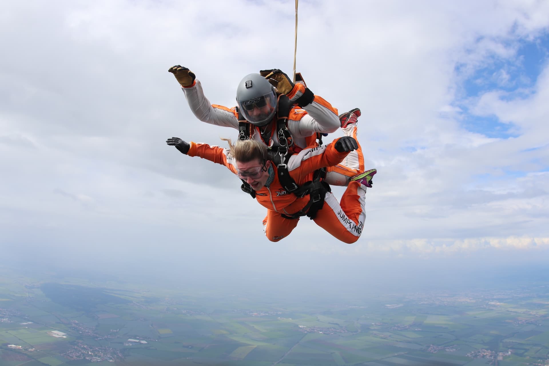 Caída libre bajo un cielo cubierto: pasajera con el pelo al viento y mono naranja e instructor tándem con casco gris, vistos de lado
