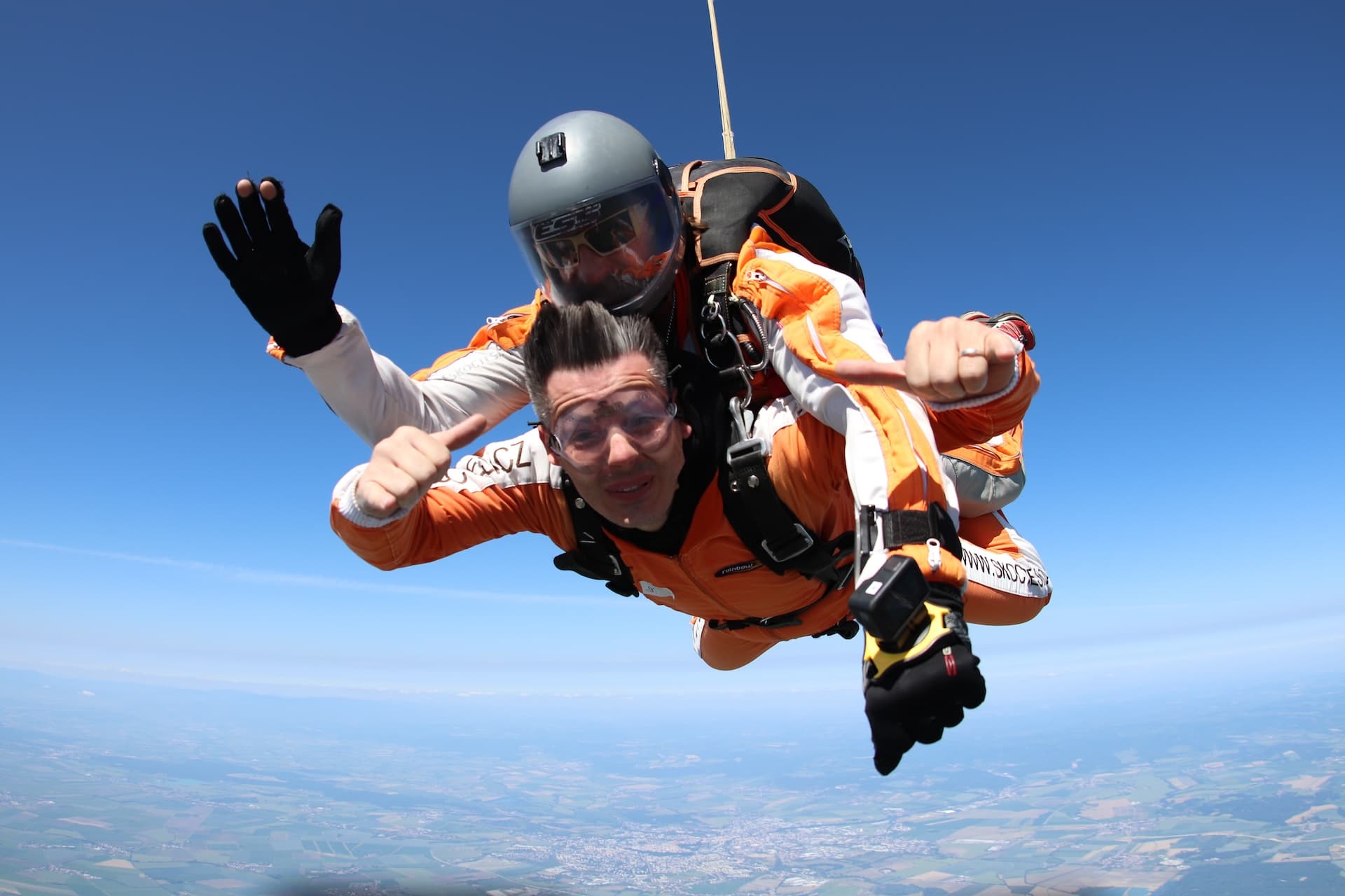 Caída libre: un pasajero con mono naranja levanta el pulgar, el instructor tándem con casco gris saluda y, muy abajo, una ciudad