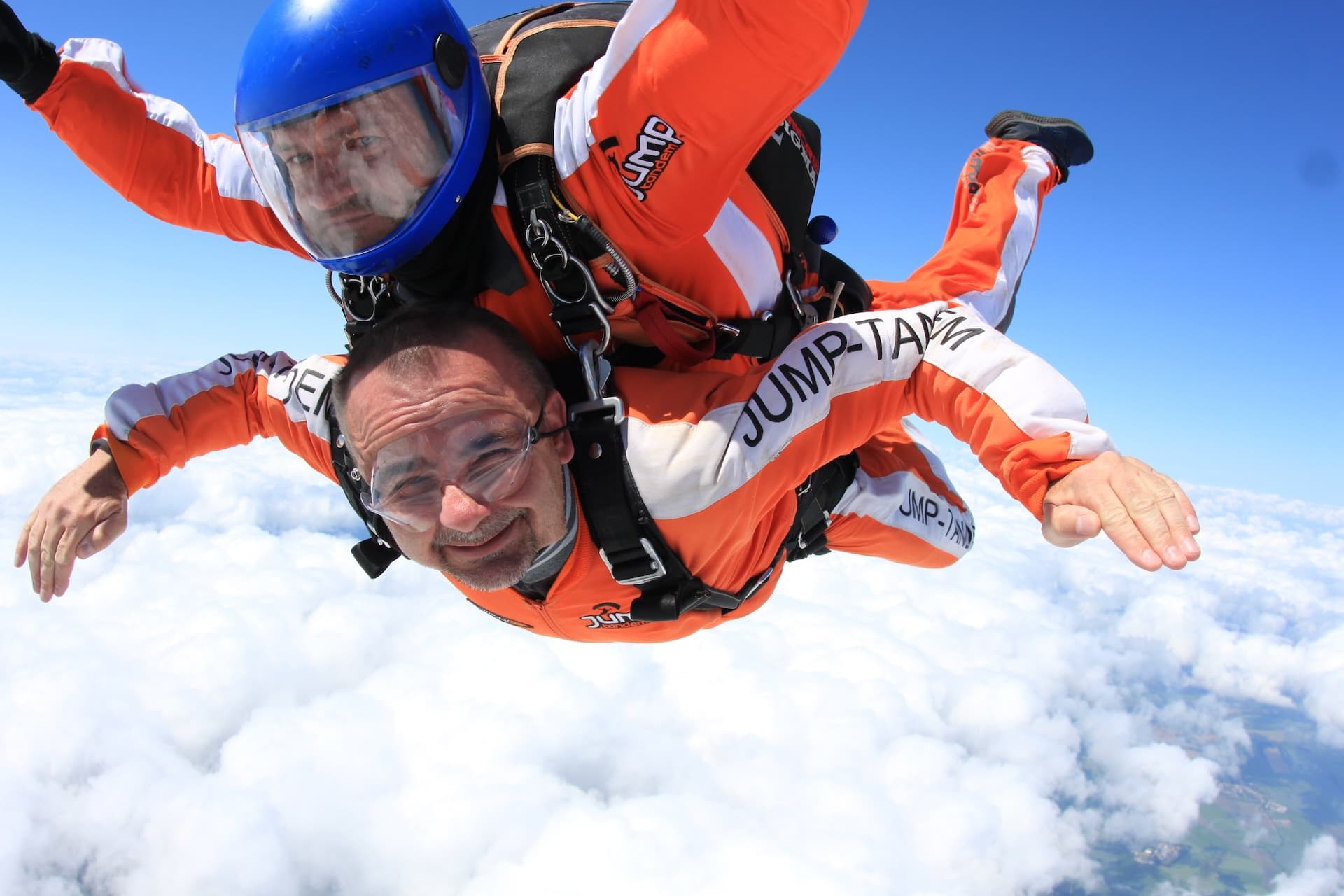 Caída libre en primer plano sobre nubes blancas: pasajero sonriente con mono naranja JUMP-TANDEM e instructor tándem con casco azul