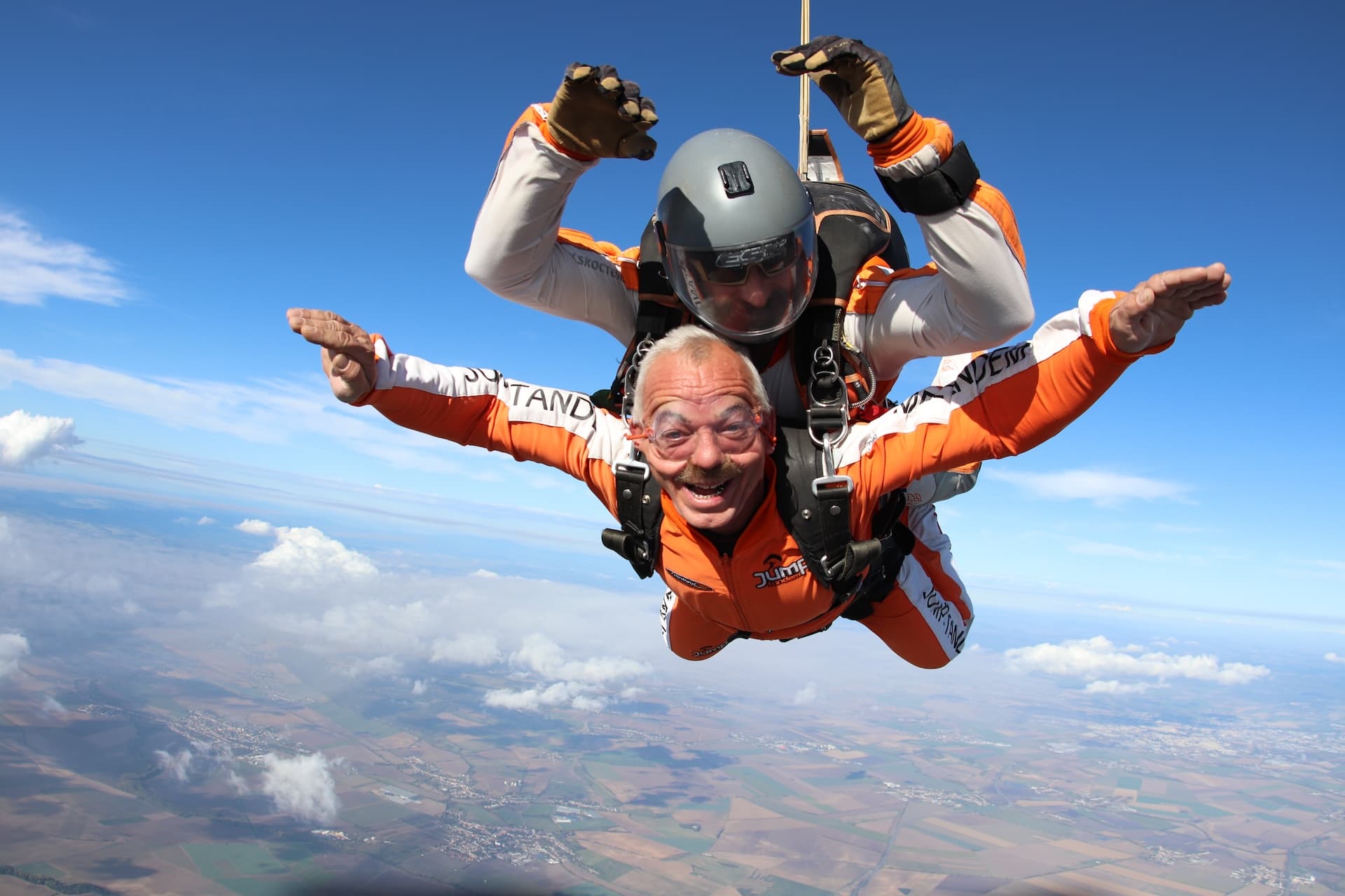 Caída libre sobre nubes dispersas y campos: pasajero con mono naranja ríe con los brazos abiertos; instructor tándem con casco gris