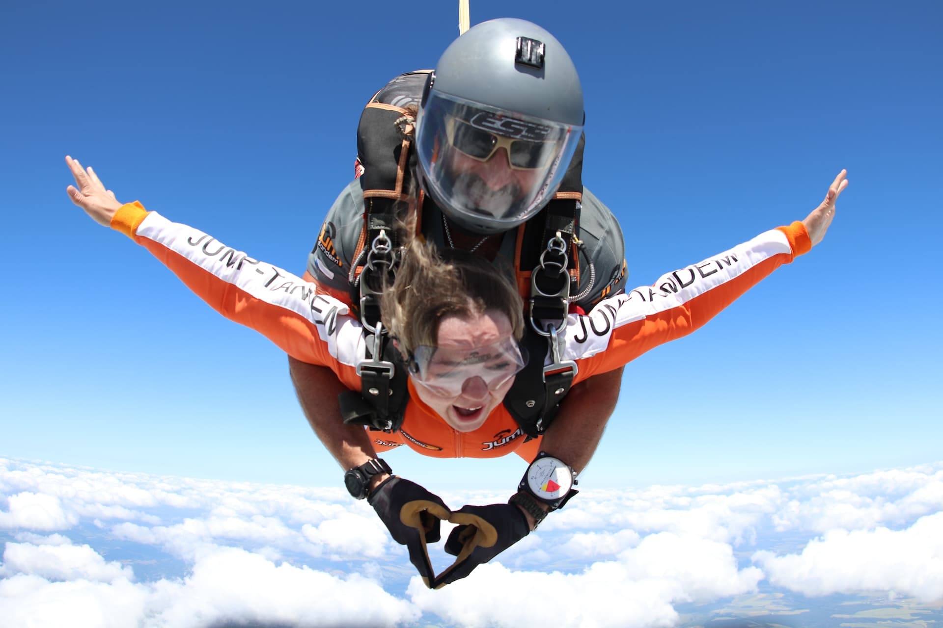 Caída libre sobre las nubes: pasajera con mono naranja JUMP-TANDEM abre los brazos; el instructor tándem forma un corazón con las manos
