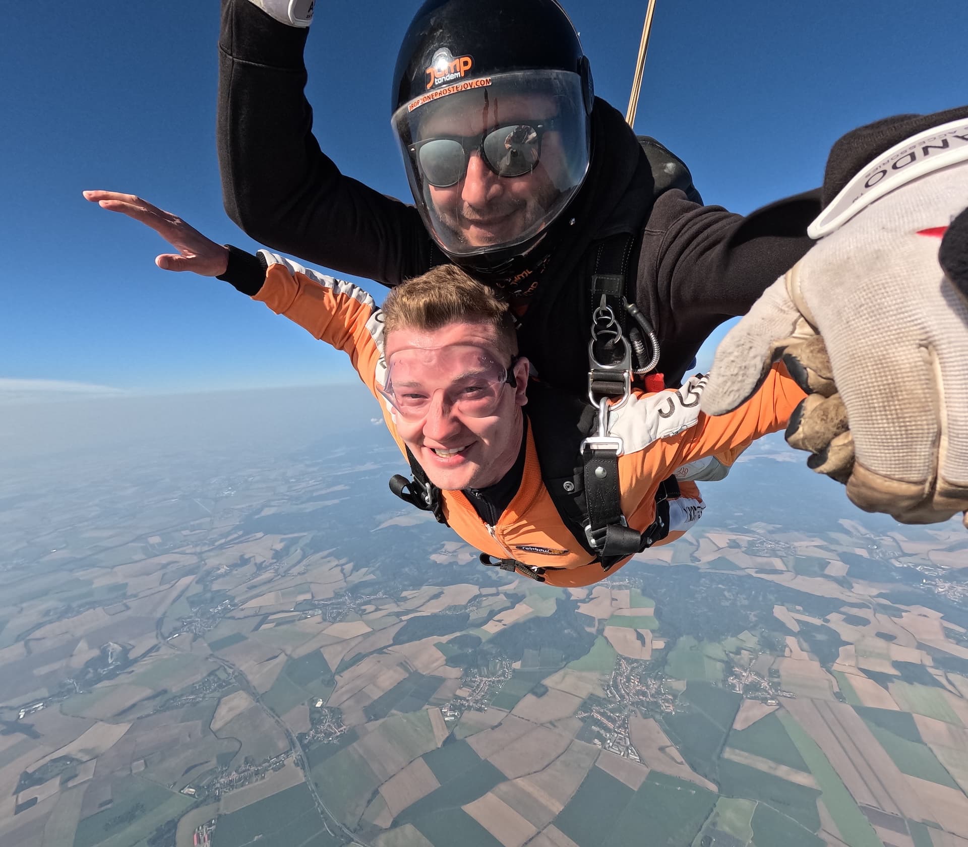 Caída libre en primer plano: pasajero sonriente con mono naranja y, detrás, el instructor tándem con casco negro Jump y gafas de sol