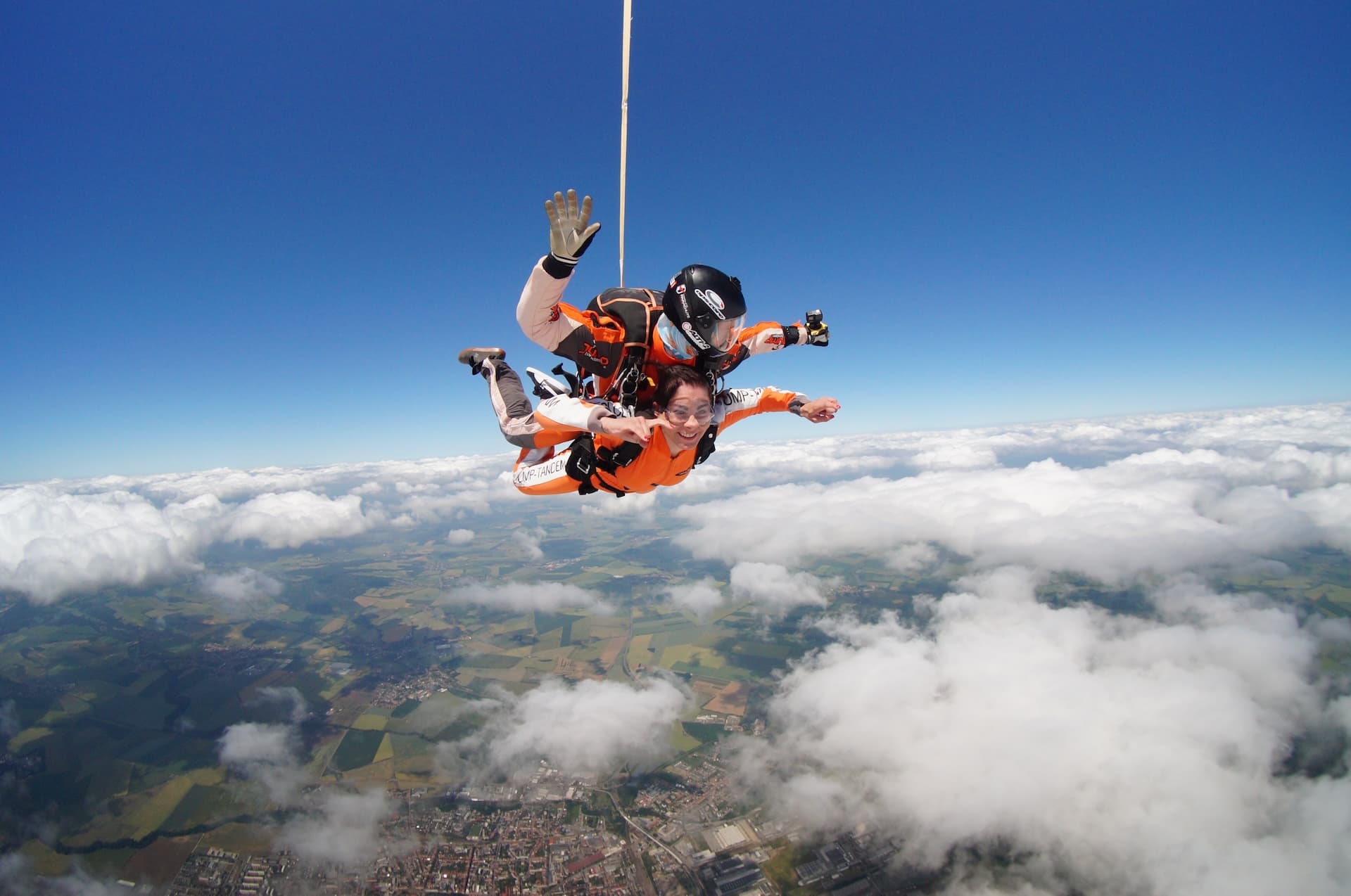 Tandem skydive 2023: tandem instructor waving and passenger in an orange jumpsuit in freefall above clouds and a town