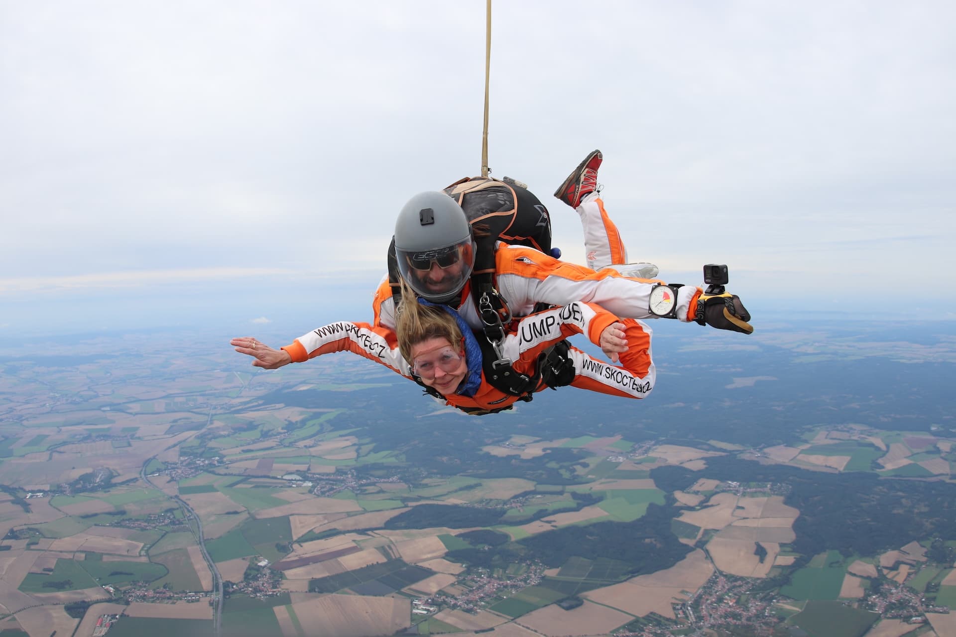 Caída libre bajo un cielo gris sobre campos y bosques: pasajera con mono naranja e instructor tándem con casco gris, vistos de lado