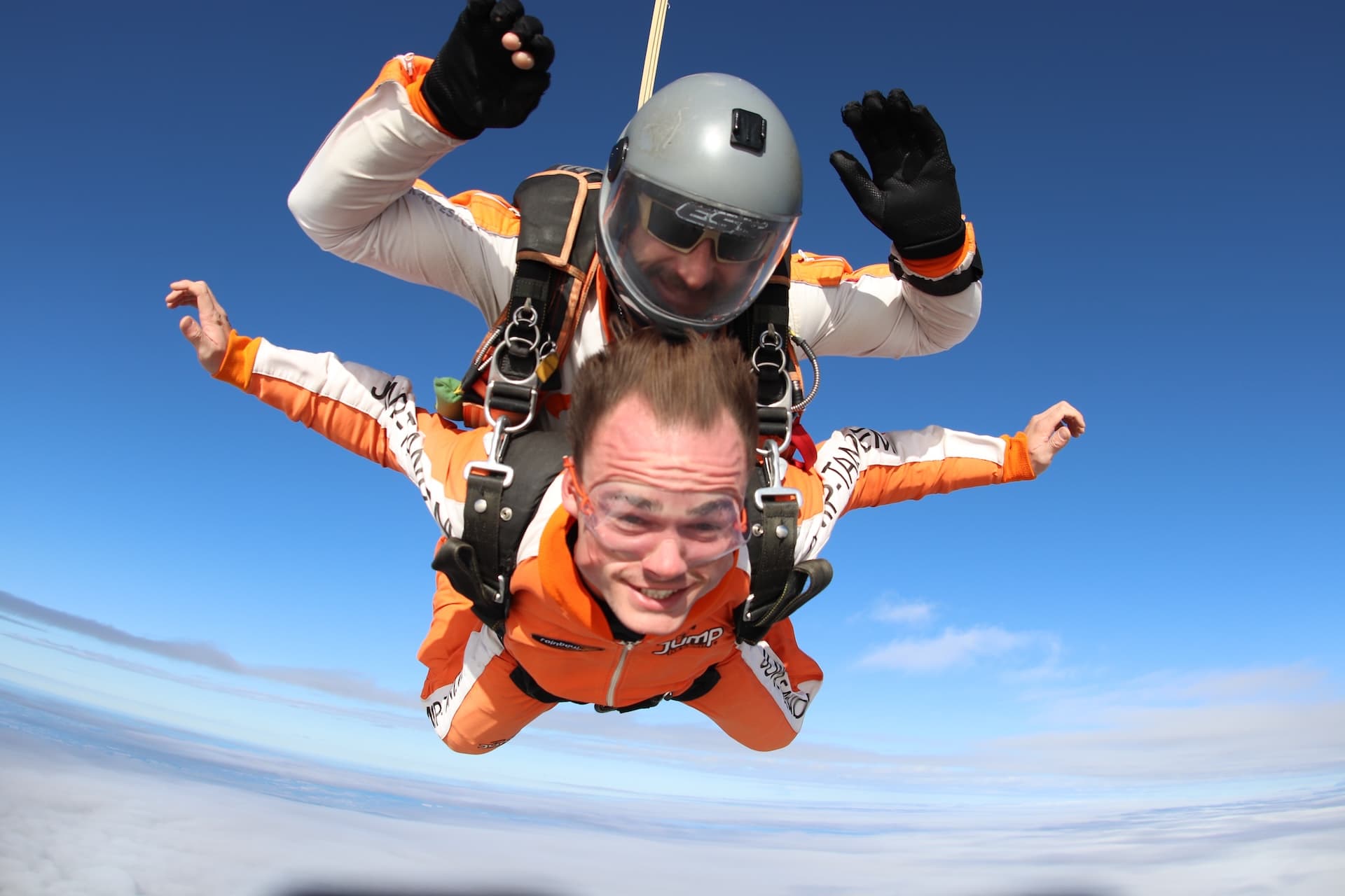 Caída libre sobre una capa de nubes: pasajero con mono naranja sonríe con los brazos abiertos; el instructor tándem saluda con ambas manos