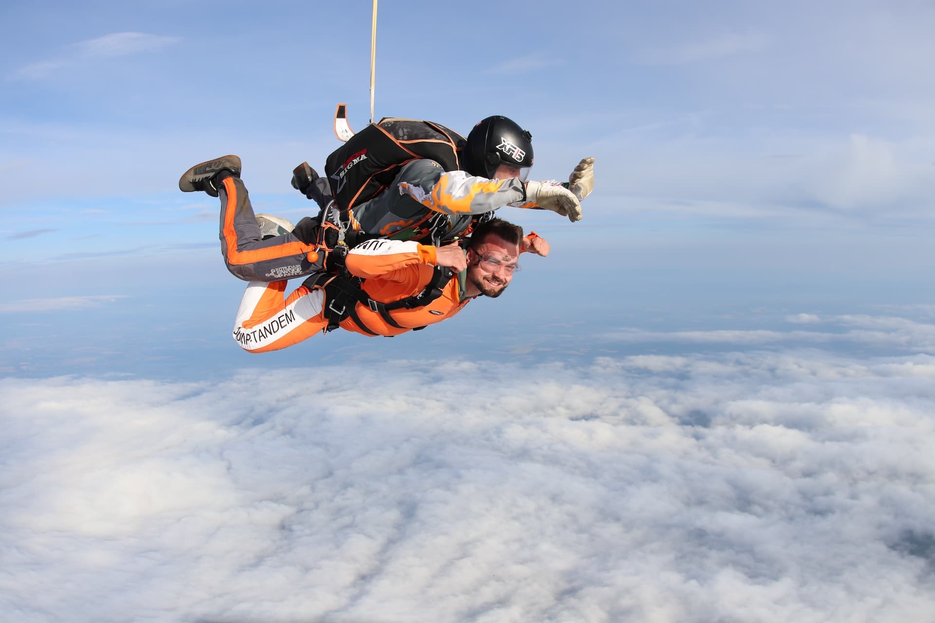 Caída libre vista de lado sobre un manto de nubes: pasajero con mono naranja JUMP-TANDEM, instructor tándem con casco negro
