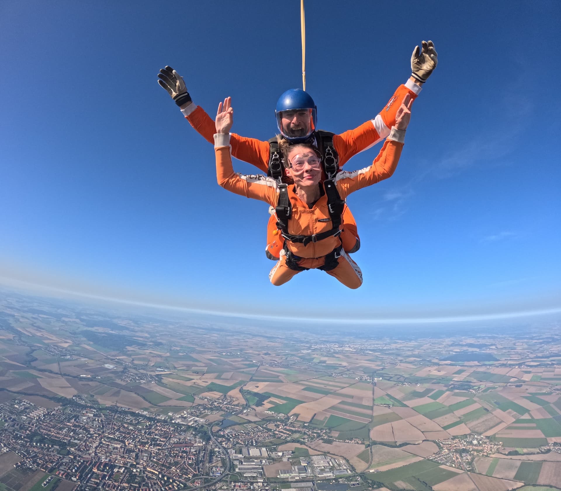 Caída libre sobre una ciudad y campos con el horizonte curvado: el pasajero y el instructor tándem con casco azul levantan los brazos