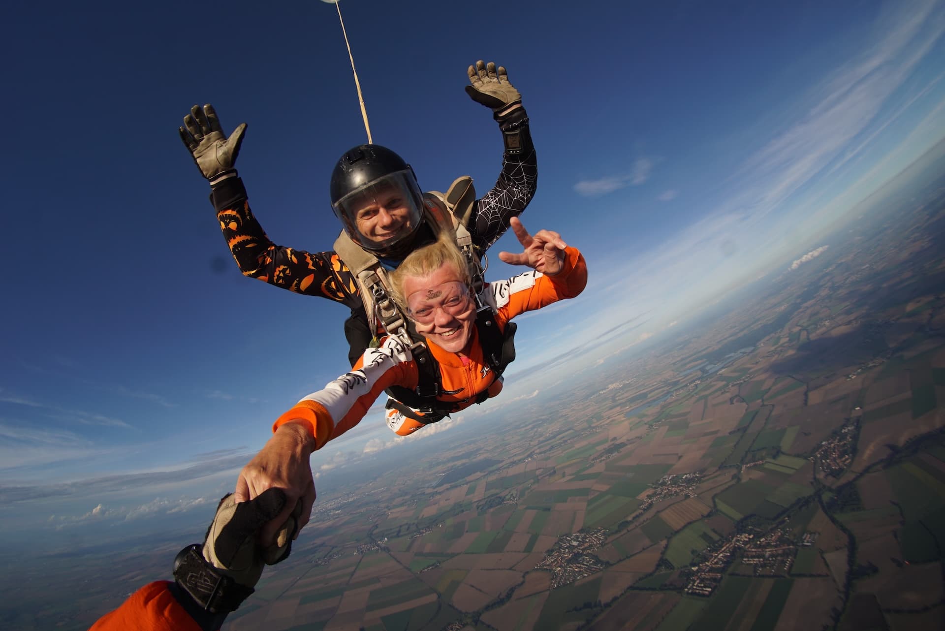 Caída libre con el horizonte inclinado: pasajera sonriente de naranja agarra la mano del cámara; el instructor tándem con casco negro saluda