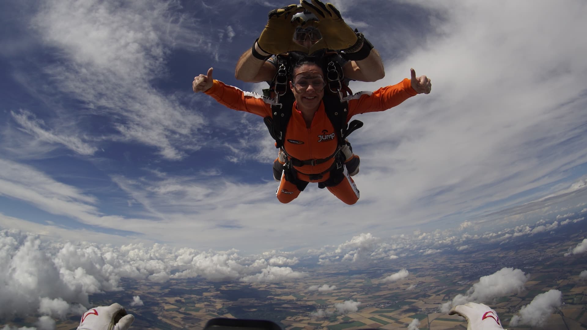 Caída libre muy por encima de las nubes: pasajero con mono naranja y los dos pulgares arriba, instructor tándem con las manos en alto