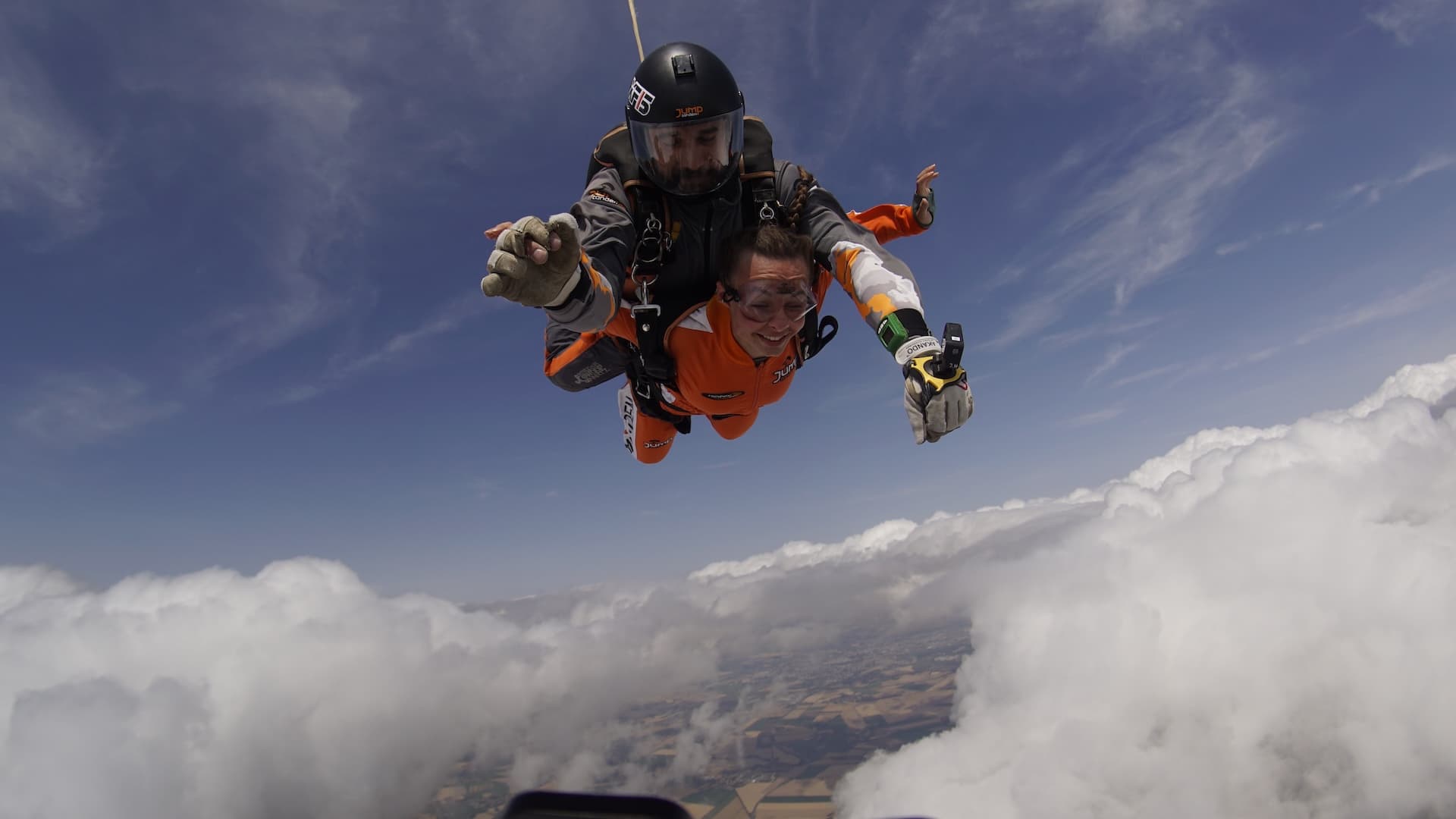 Caída libre junto a grandes nubes blancas: pasajero sonriente con mono naranja e instructor tándem con casco negro, brazos abiertos