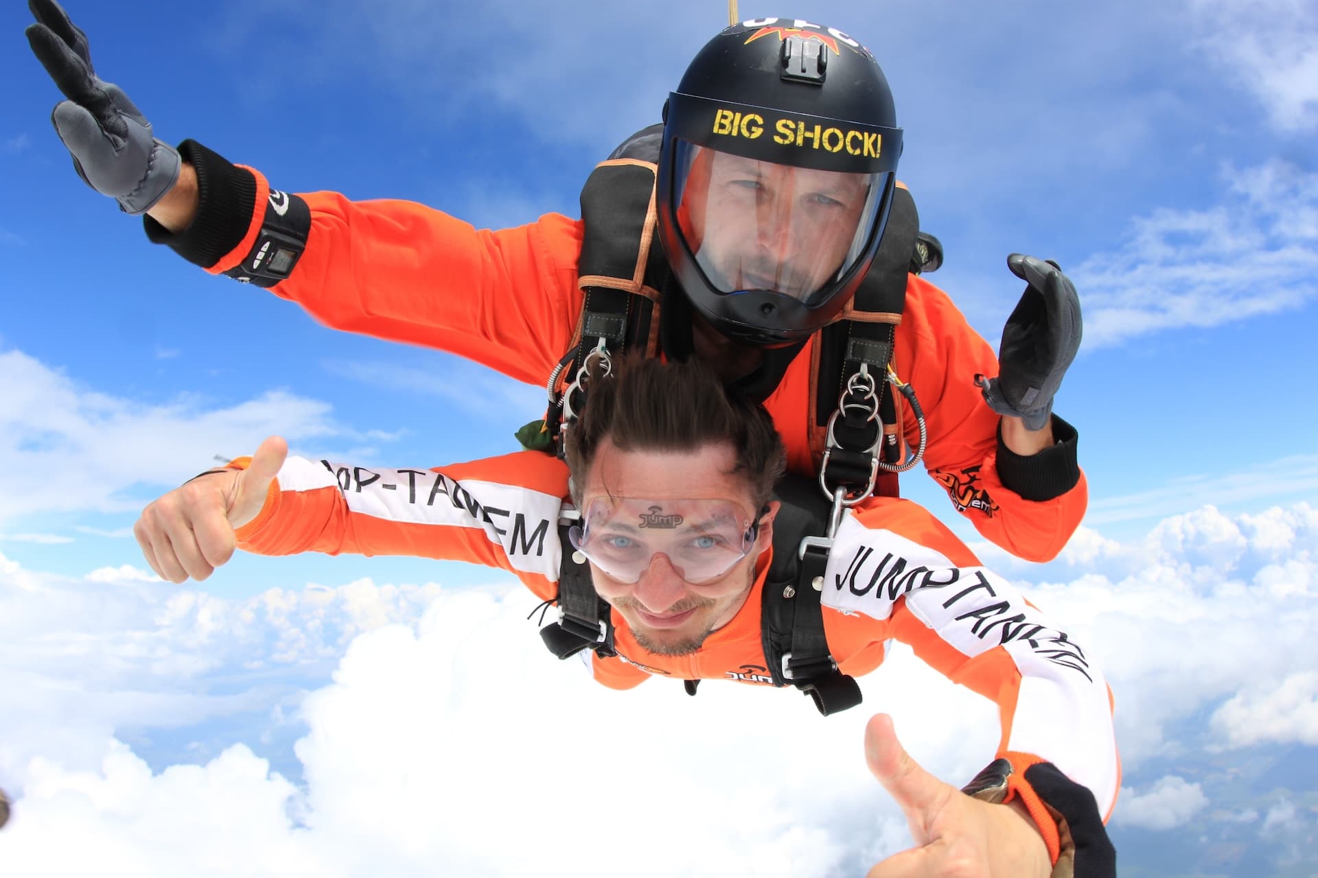 Caída libre sobre las nubes: pasajero con mono naranja y los dos pulgares arriba; el casco negro del instructor tándem dice Big Shock!