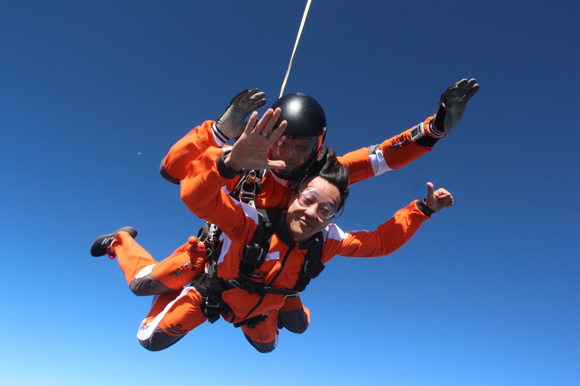 Caída libre en un cielo azul intenso: pasajero con mono naranja saluda a la cámara con el pulgar arriba; instructor tándem con casco negro