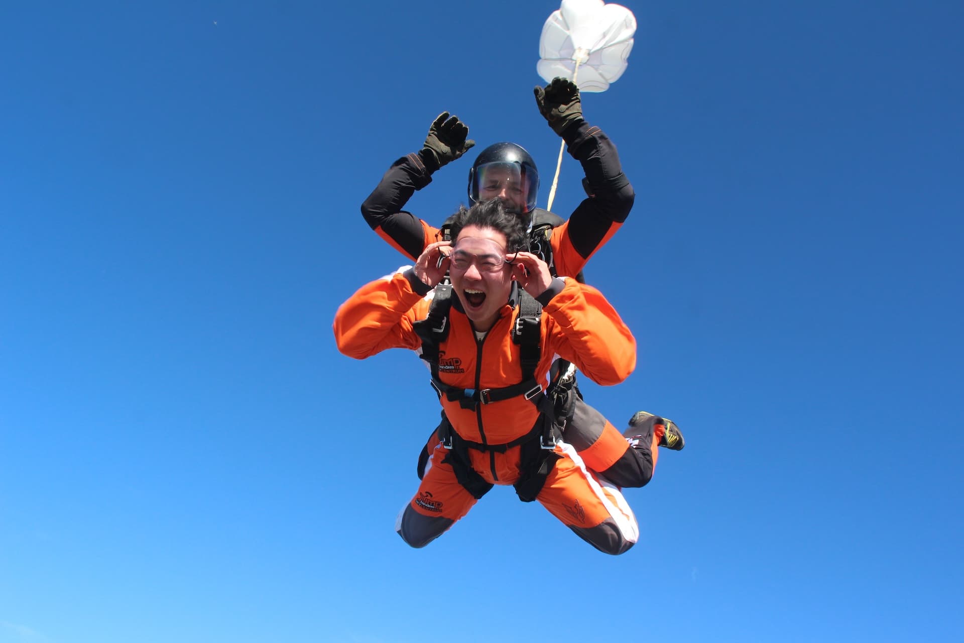 Caída libre: pasajero con mono naranja grita sujetándose las gafas; instructor tándem con los brazos en alto y estabilizador blanco encima