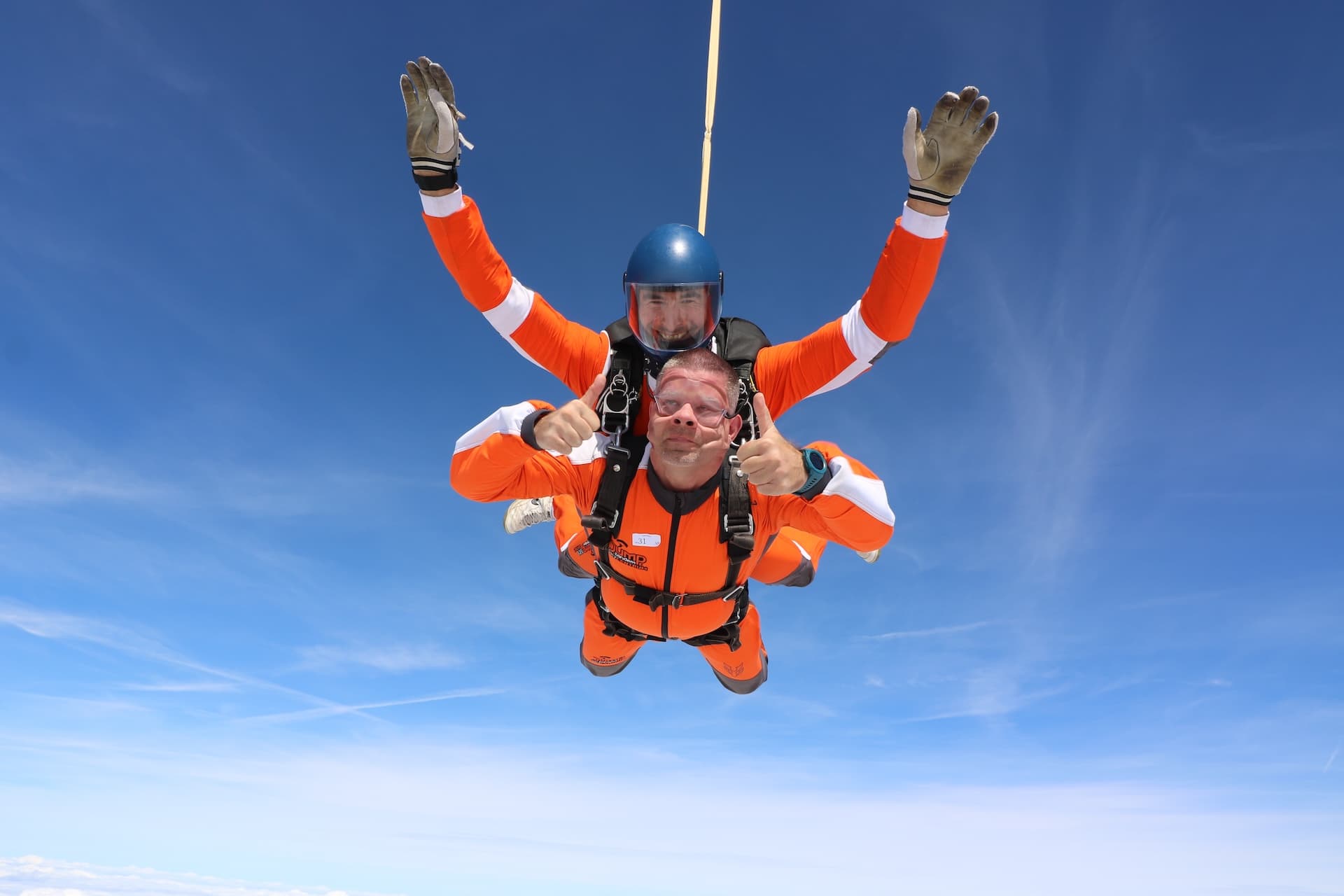 Caída libre: pasajero con mono naranja y los dos pulgares arriba; el instructor tándem con casco azul estira los brazos hacia arriba