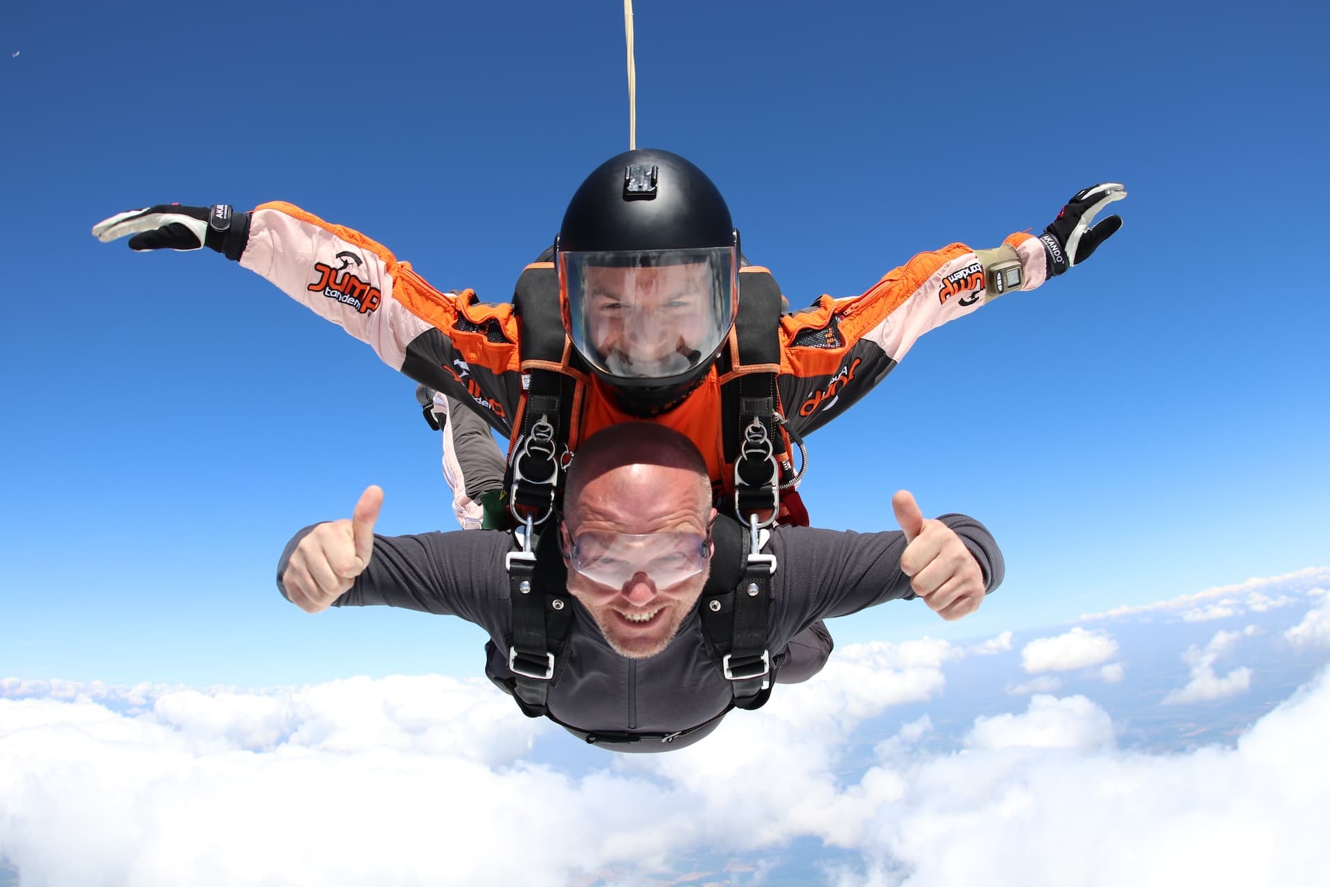 Caída libre sobre nubes blancas: pasajero de gris con los dos pulgares arriba; instructor tándem con mono Jump blanco y naranja