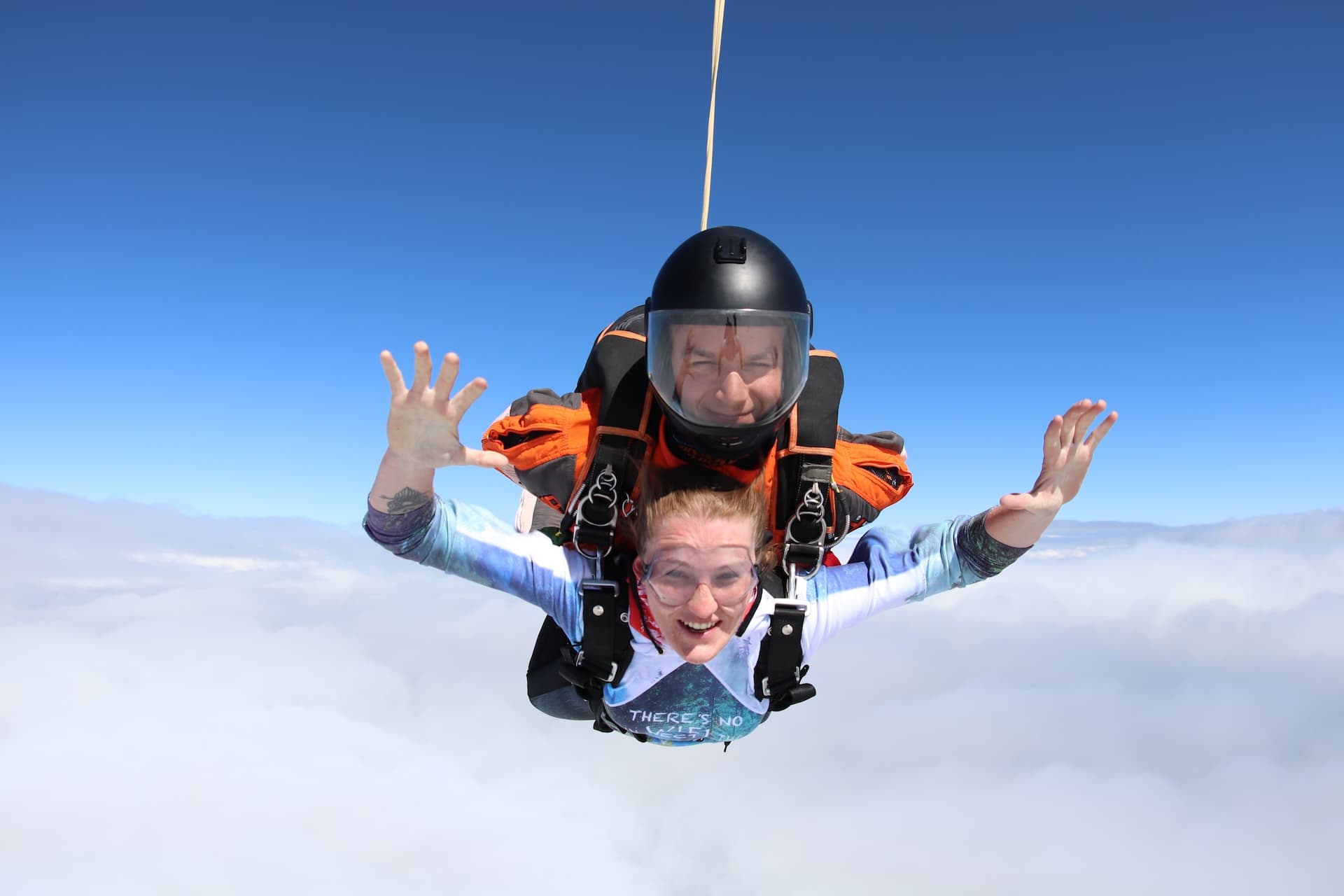 Caída libre sobre una capa de nubes: pasajera sonriente con camiseta azul claro y los dedos abiertos; instructor tándem con casco negro