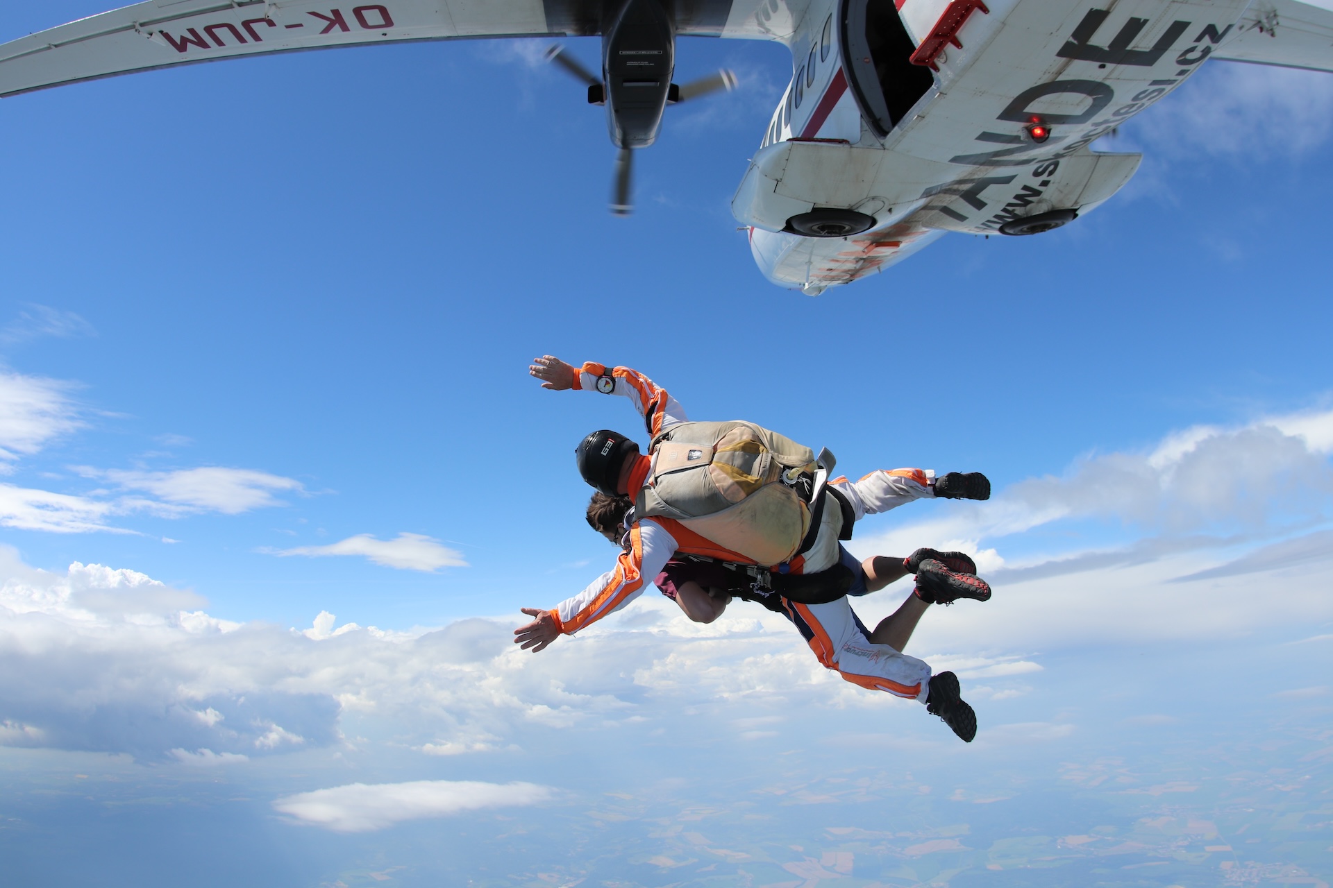 Instructor tándem y pasajero justo tras el salto, vistos desde arriba: sobre ellos el avión blanco y rojo, debajo las nubes