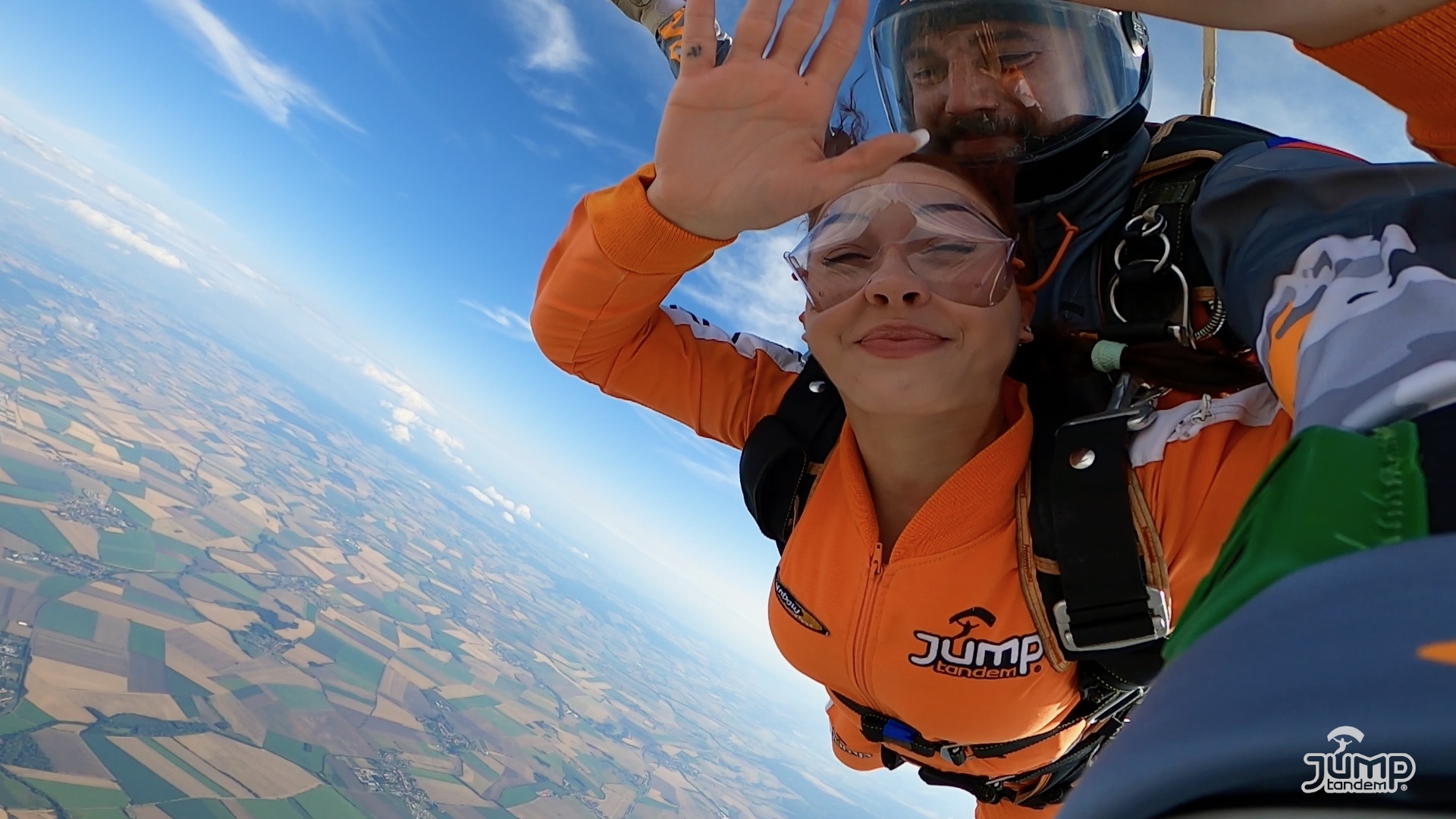 Tandem jumps Prostějov 2024: passenger in an orange JUMP-TANDEM jumpsuit waving at the camera in freefall above the fields