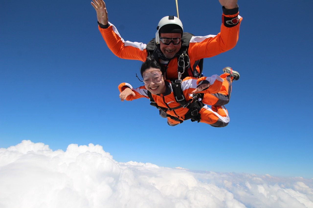 Tandem jumps Příbram 2024: tandem instructor in a white helmet and smiling passenger in orange in freefall above clouds