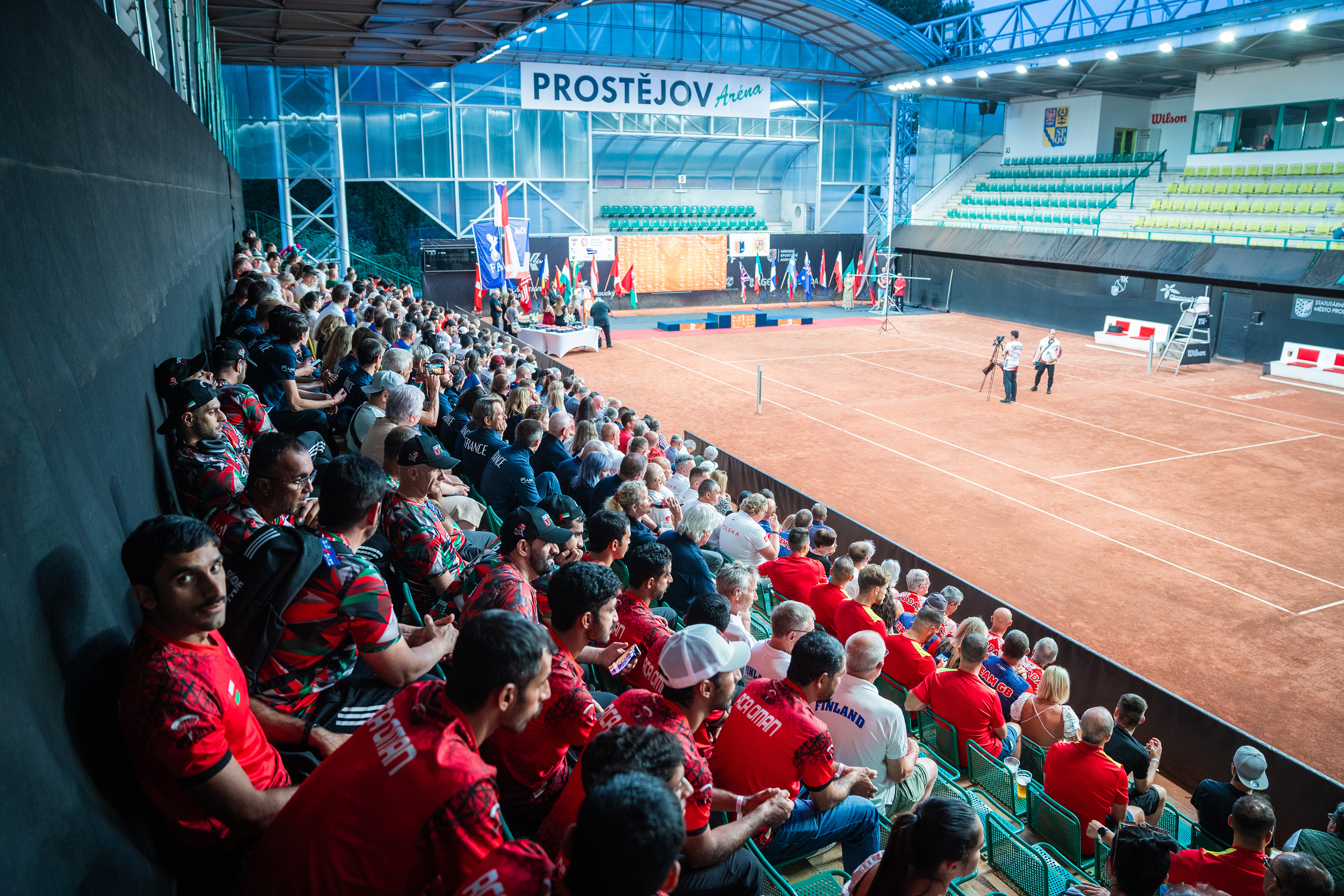 Campeonatos del Mundo FAI 2024: equipos en la grada del Prostějov Arena junto a una pista de tierra batida y un escenario con banderas