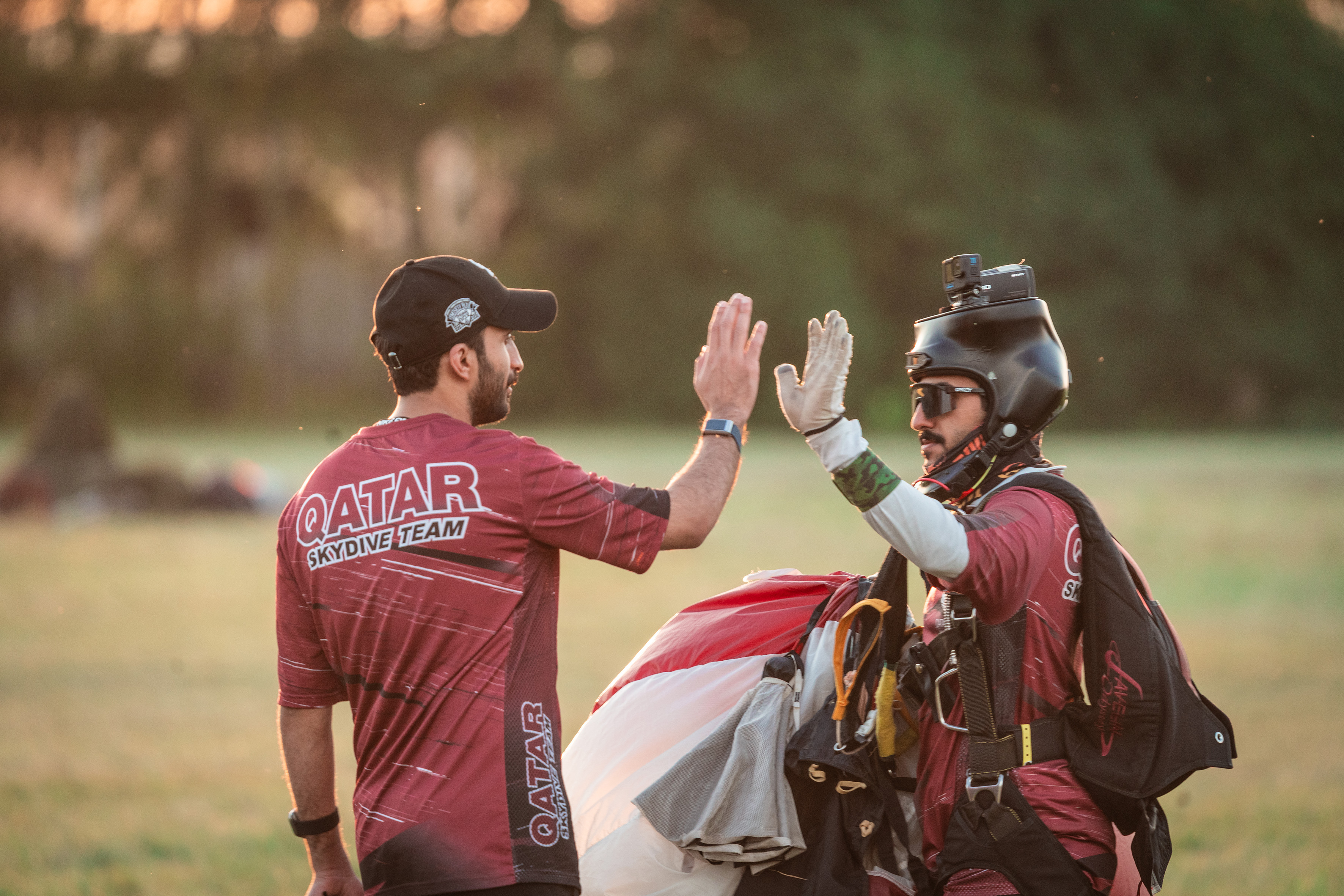 Campeonatos del Mundo FAI 2024: dos miembros del Qatar Skydive Team chocan las manos, uno con casco con cámara y el paracaídas en brazos